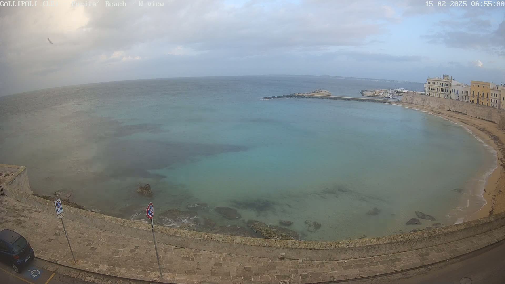 A mostly cloudy day overlooks a calm, teal-colored bay with a sandy beach and buildings along the shore.
