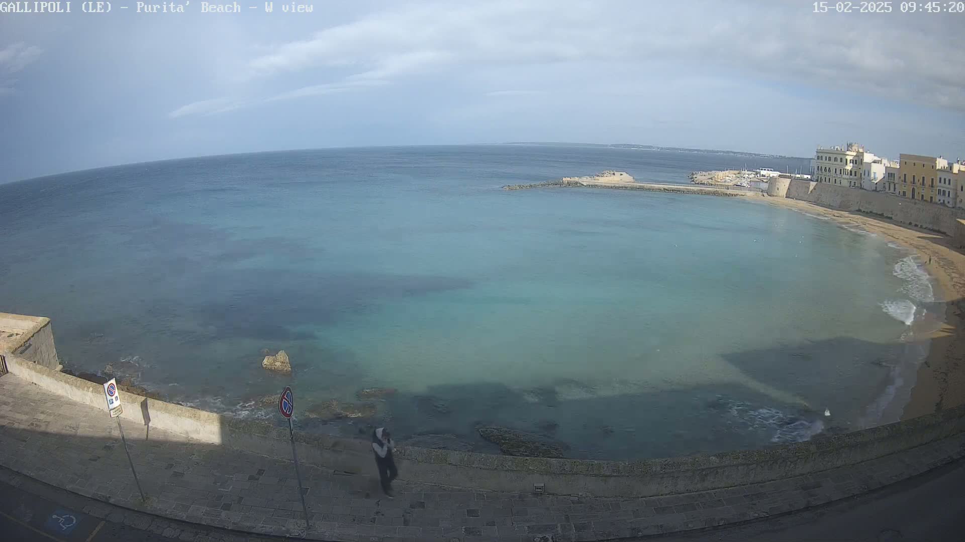 A partly sunny day shows a calm, teal-colored bay with a sandy beach curving along a stone wall and buildings.