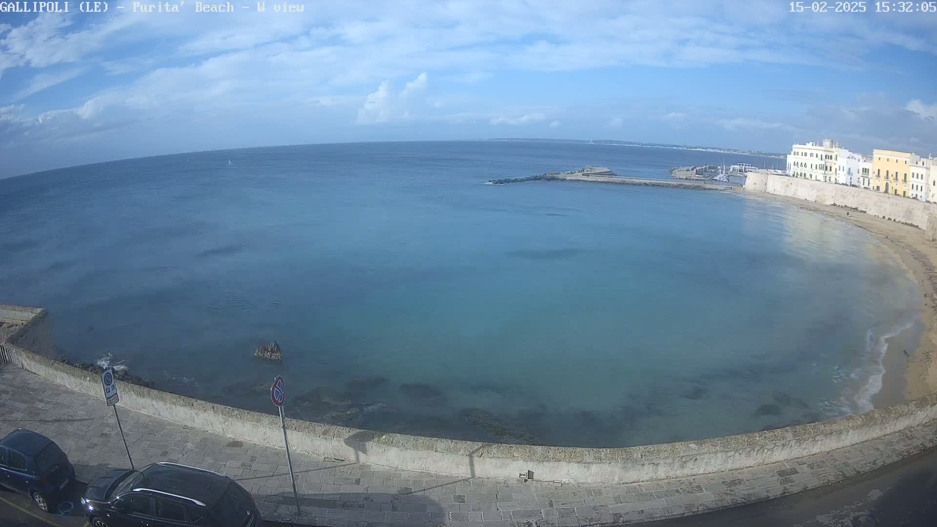A mostly sunny day reveals a calm, teal-colored bay with a sandy beach and stone buildings along the shoreline.