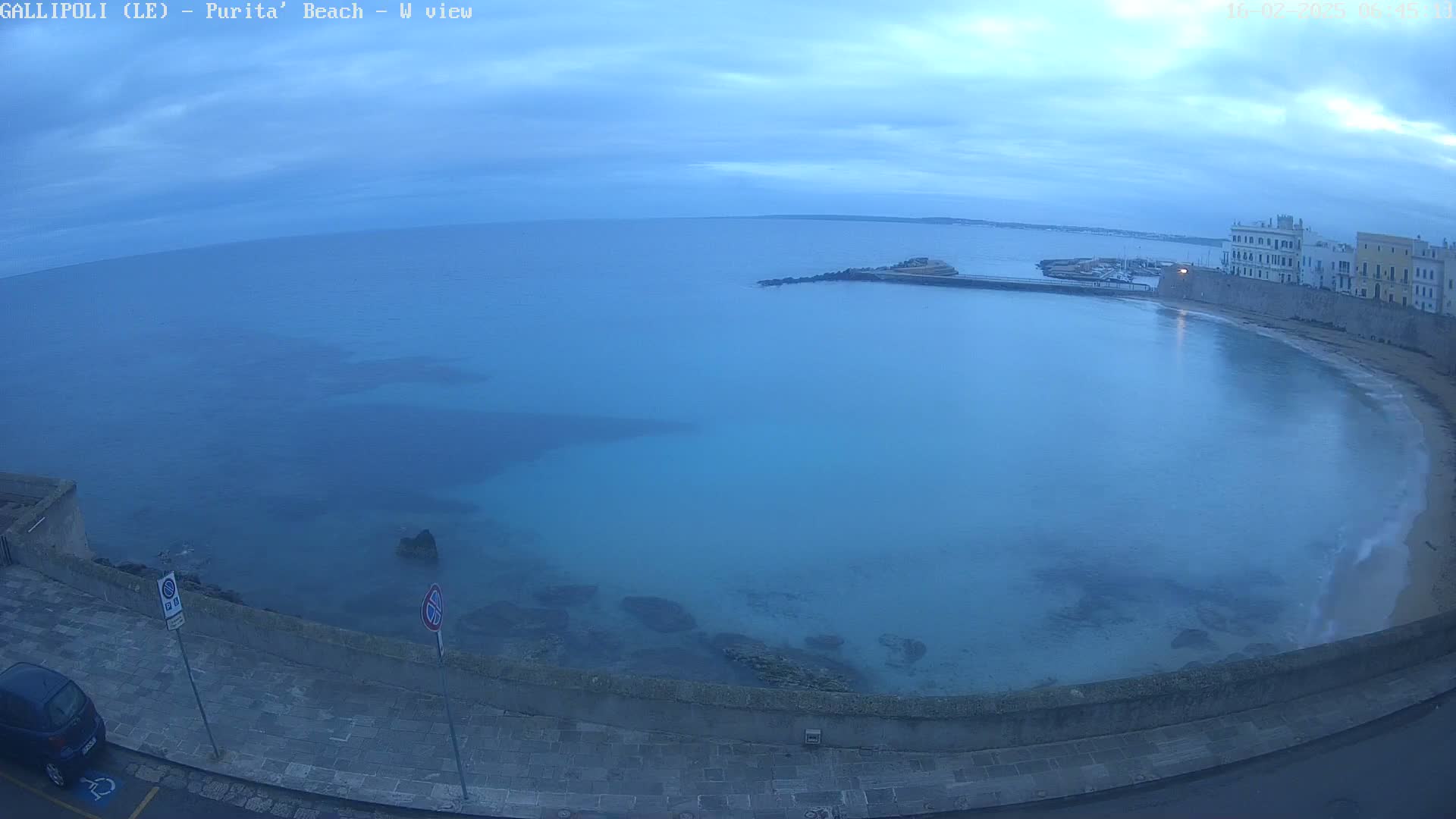A calm, blue sea curves gently around a sandy beach and stone buildings under a mostly cloudy, dusky sky.
