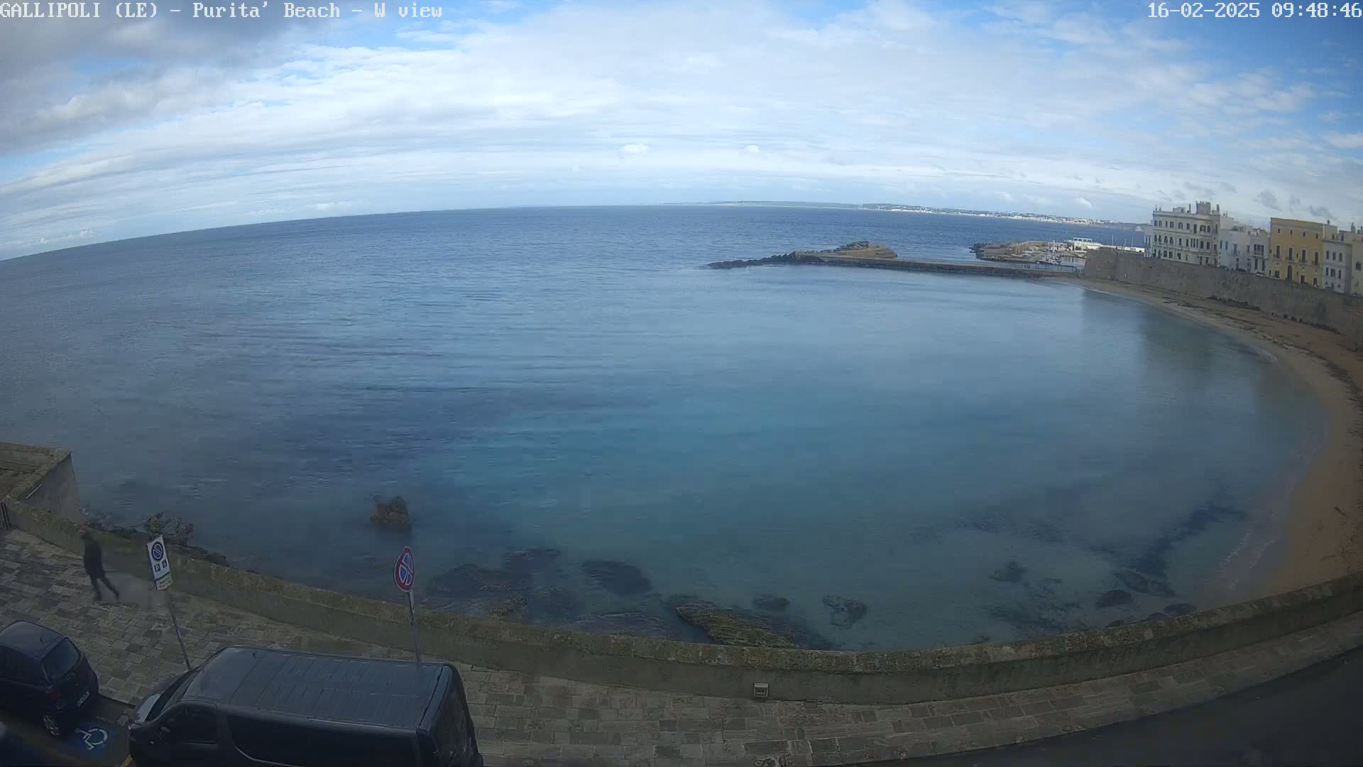 A partly cloudy day reveals a tranquil bay with calm, teal water lapping a sandy shore, backed by a stone wall and buildings.
