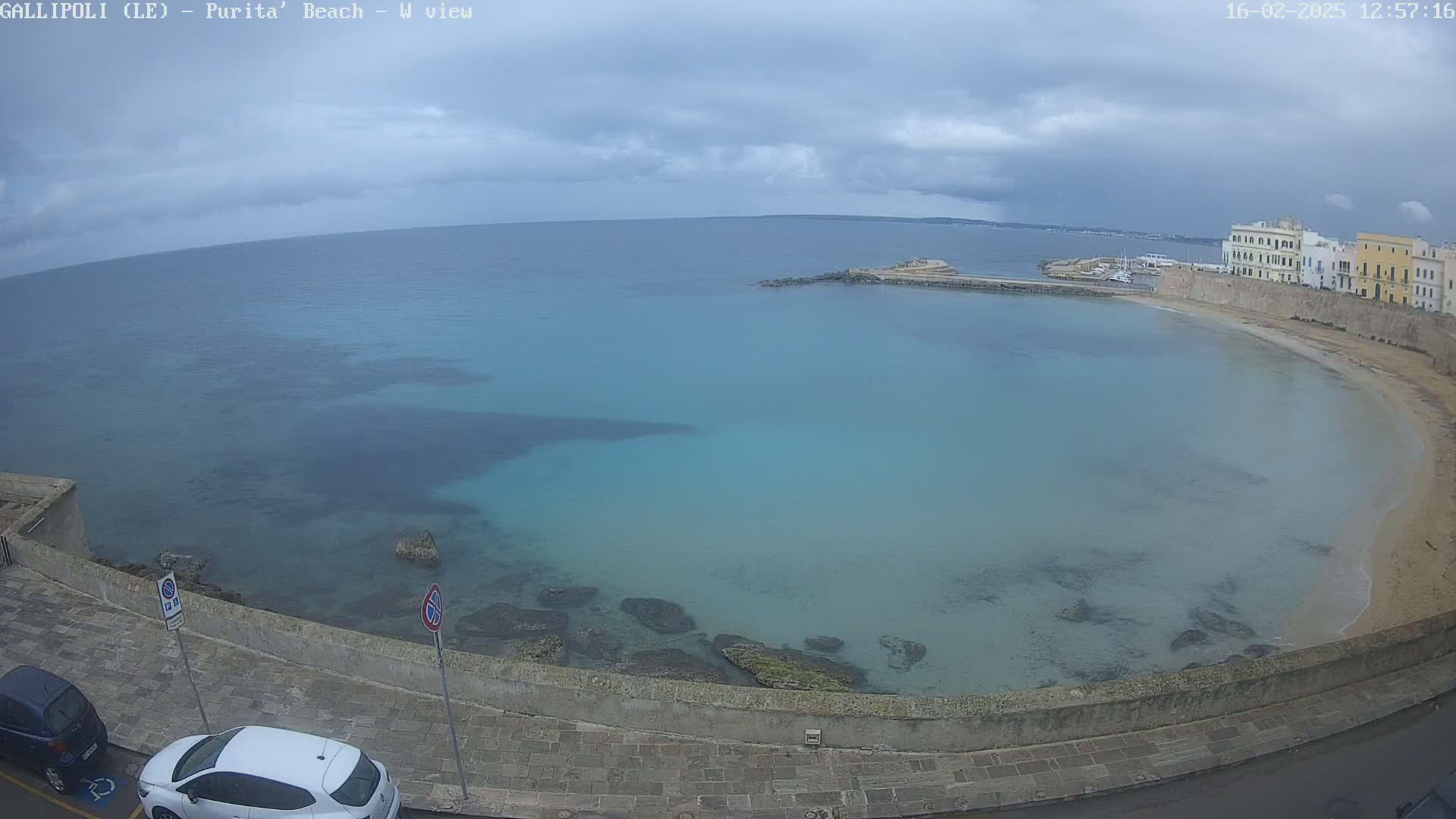 A partly cloudy day reveals a tranquil, turquoise bay with a sandy beach and stone buildings, viewed from a high vantage point above a paved road.