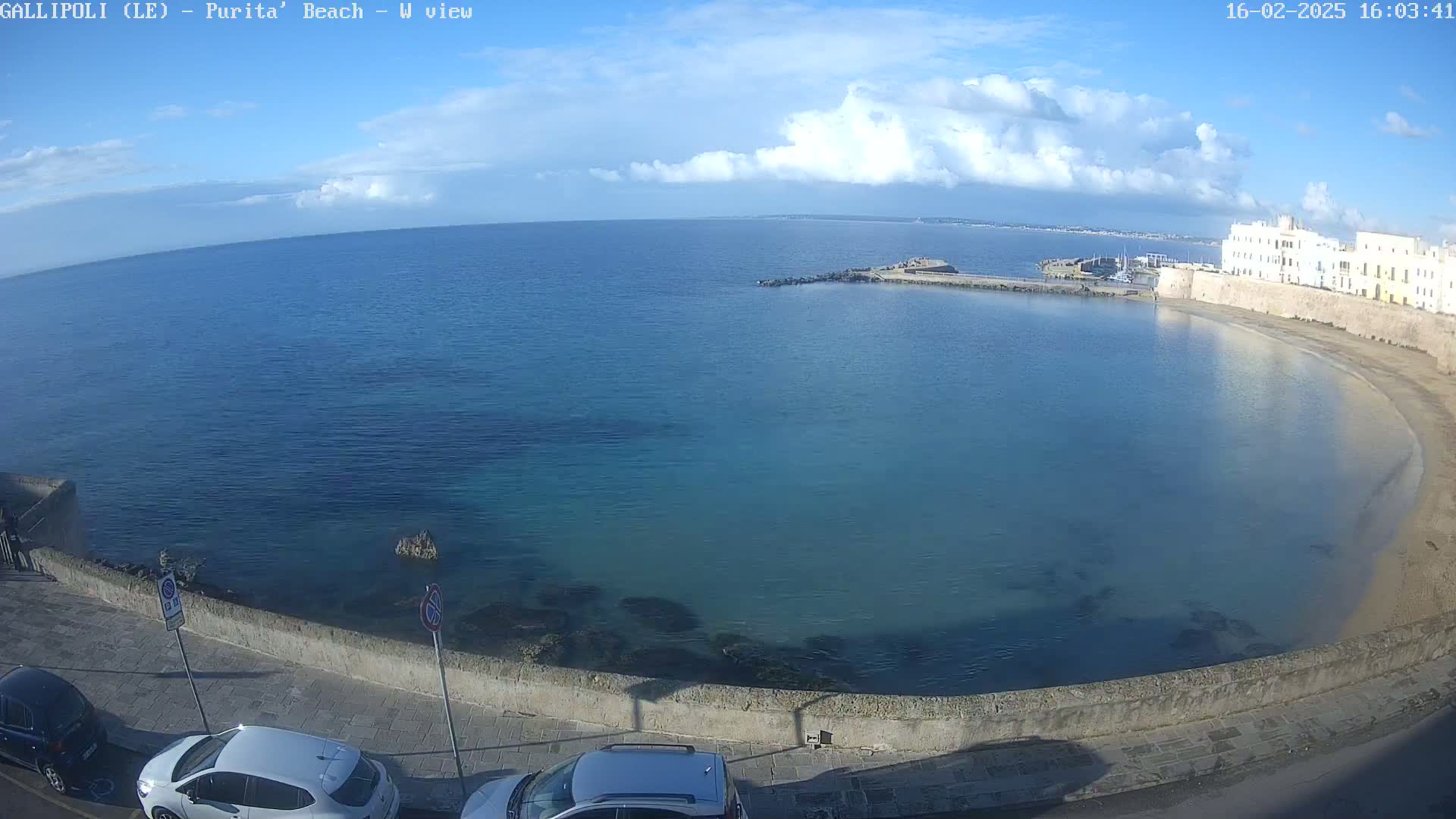 A partly cloudy day shows a tranquil bay with clear, teal water, a sandy beach, and a stone wall bordering a street with parked cars.