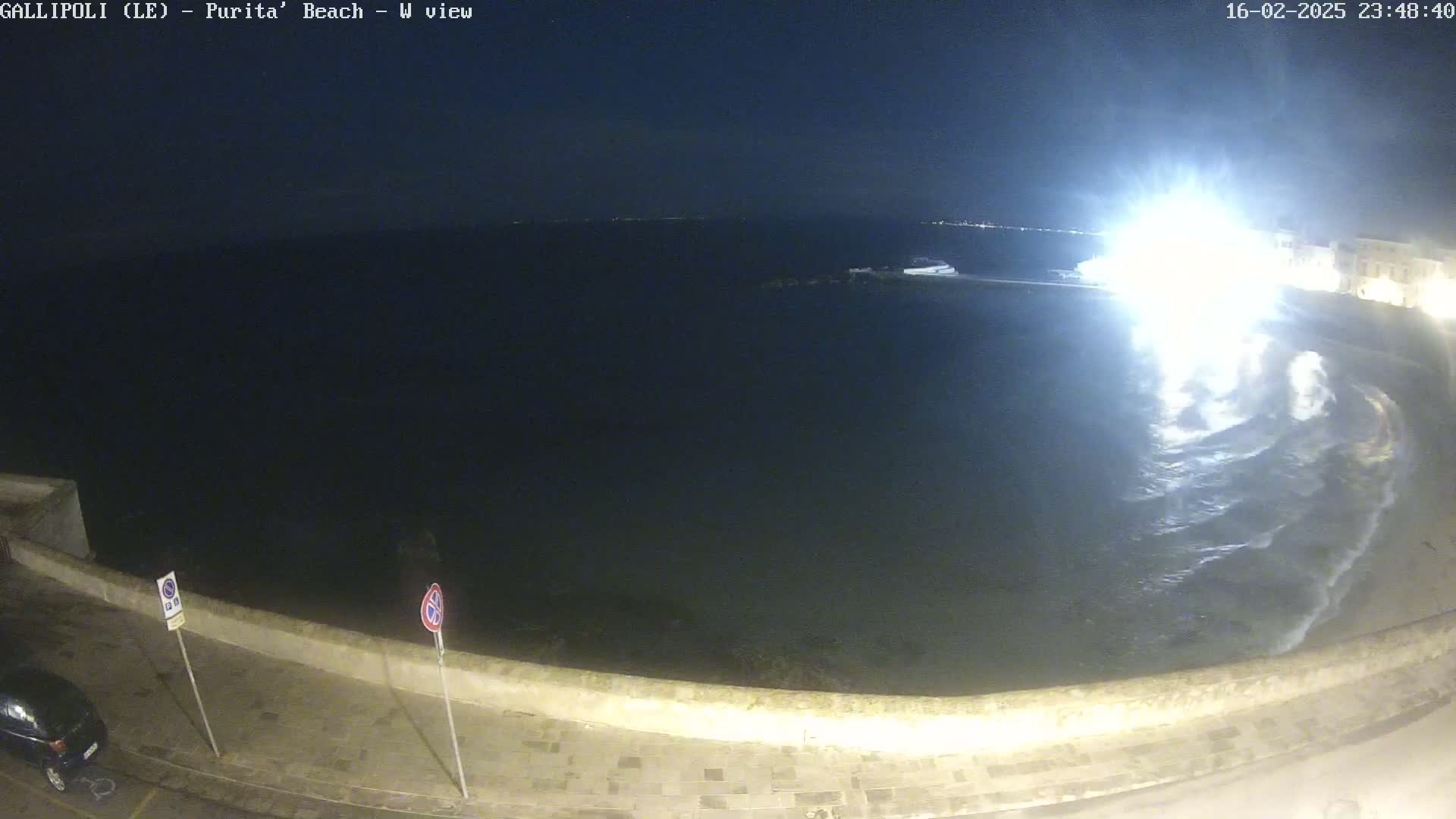 A nighttime, seaside view shows dark water with small waves lapping against a low stone wall, illuminated by bright lights.