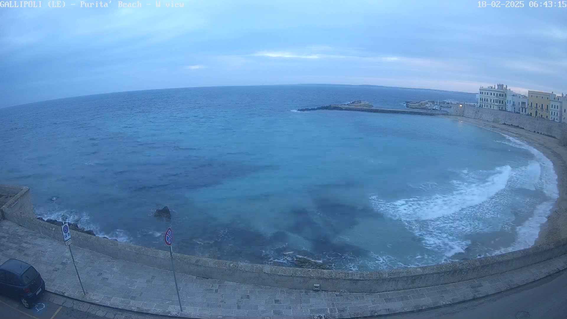 A high-angle view shows a calm, blue-green sea lapping gently against a stone wall next to a paved road with a car parked, under an overcast sky.