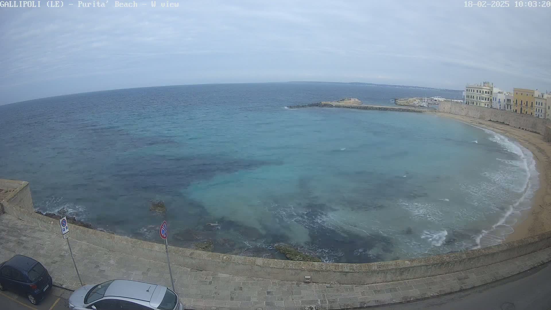 A partly cloudy day shows a view of a calm, teal-colored bay with a sandy beach curving along a stone wall, next to buildings, with two cars parked along the wall.