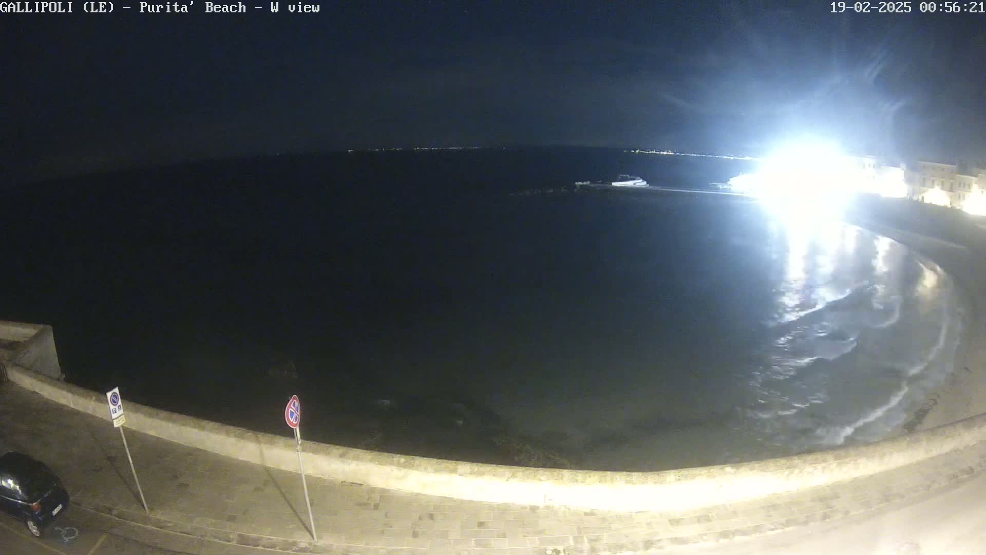 A nighttime view of a calm, dark ocean lapping gently at a stone wall along a paved shore, illuminated by a bright light source.