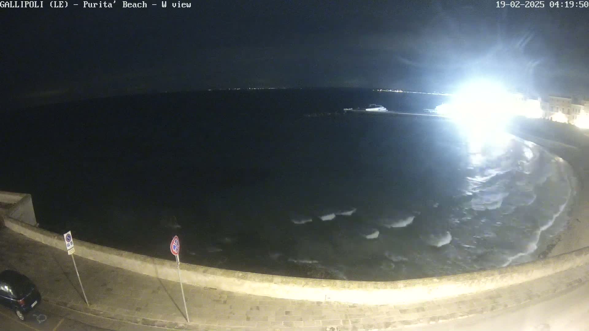 A nighttime view of a dark, wavy sea lapping at a stone wall, with small waves visible near the shore under a dark sky.