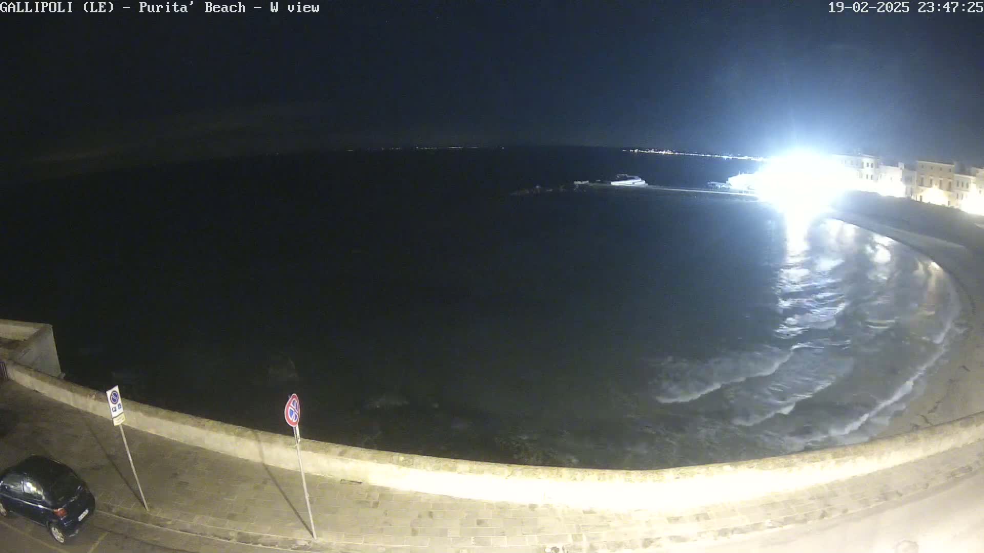 A nighttime, overhead view shows a calm, dark-colored sea lapping gently at a crescent-shaped shore, with a low wall separating the water from a paved area.