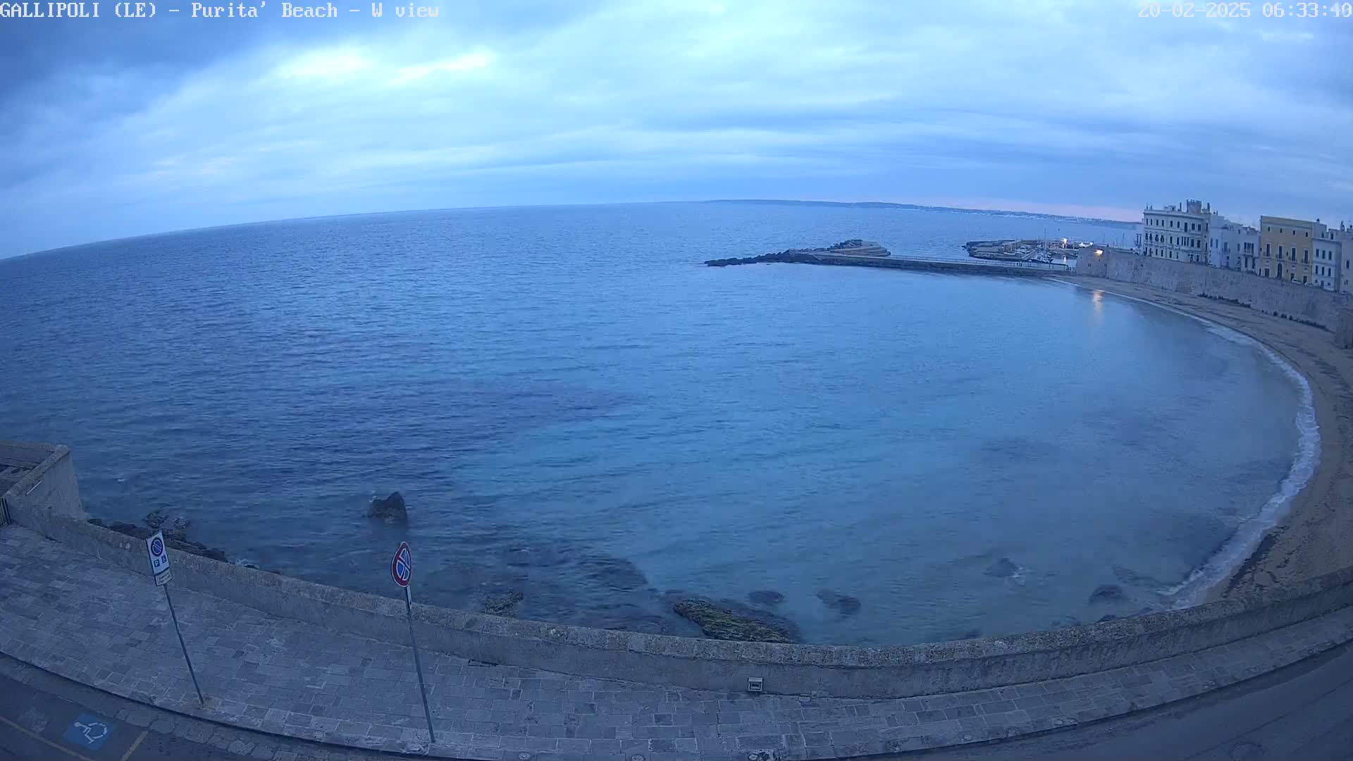 A mostly clear, dusky sky overlooks a calm, blue-green sea curving around a stone shoreline and old buildings.
