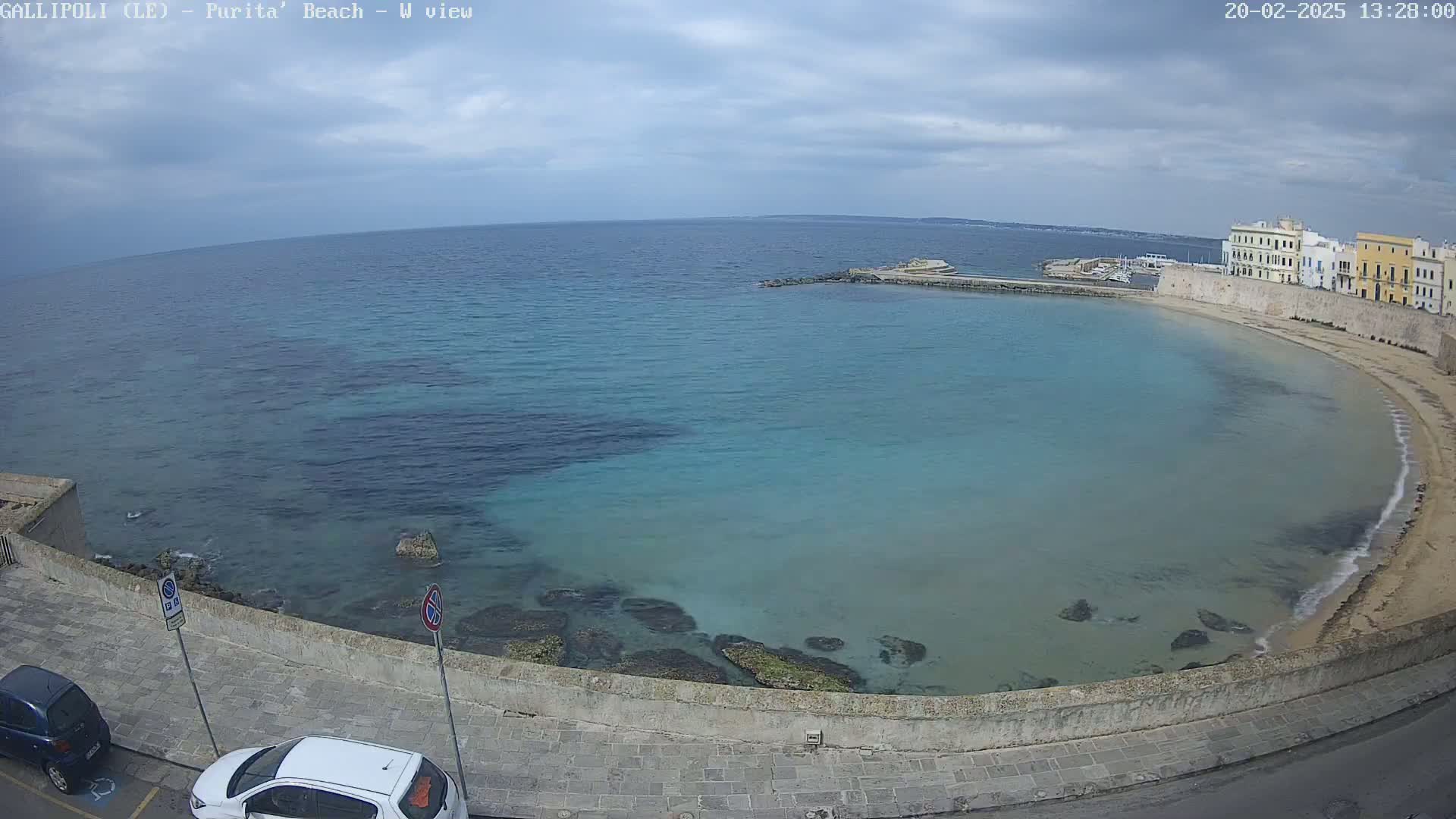 A partly cloudy day overlooks a calm, turquoise bay curving along a sandy beach and stone buildings.