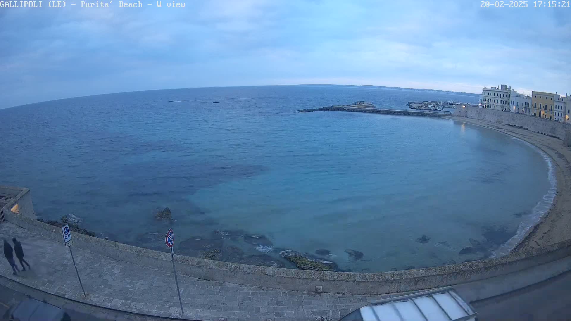 A curving bay with calm, clear blue water laps a sandy beach next to a stone wall and buildings under a partly cloudy sky.