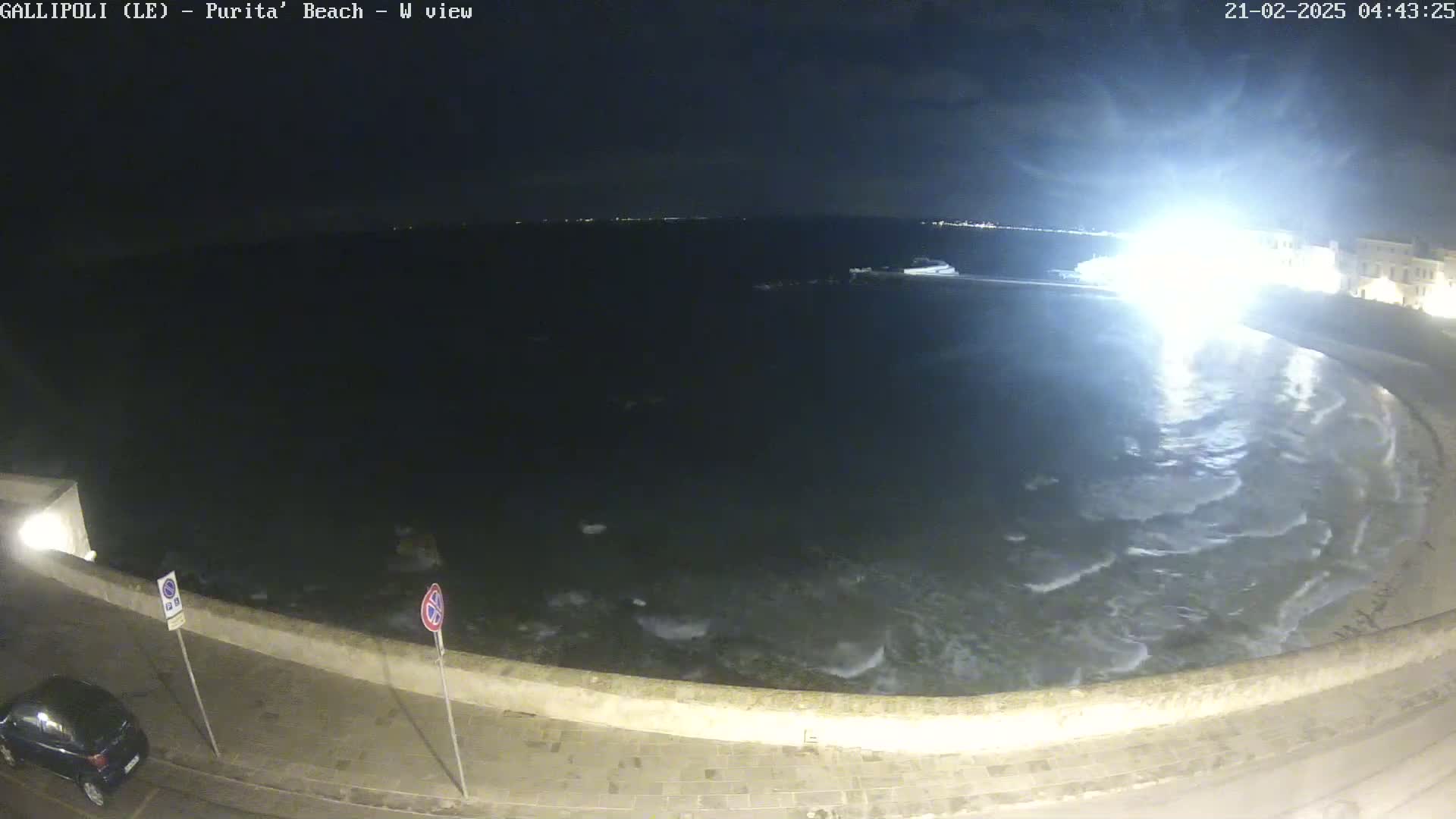 A nighttime view of a beach with gentle waves lapping against a stone wall, under a dark sky.