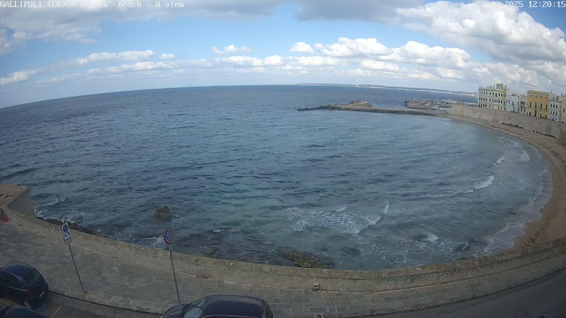 A partly cloudy day shows a coastal scene with a calm sea lapping a sandy beach next to a stone wall and buildings.