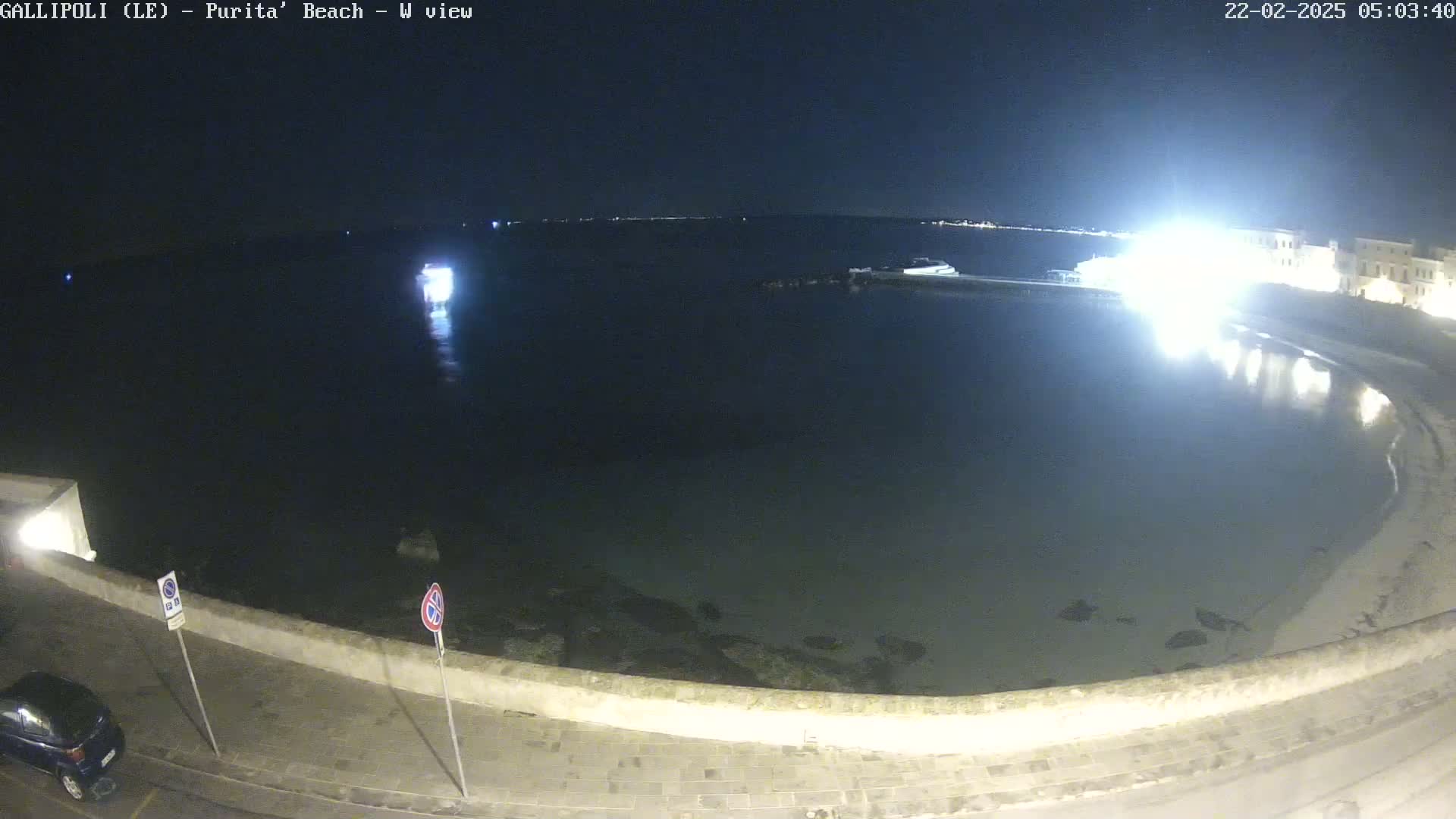 A nighttime view of a calm bay, illuminated by artificial lights, with a shoreline featuring a low wall and a parked car.