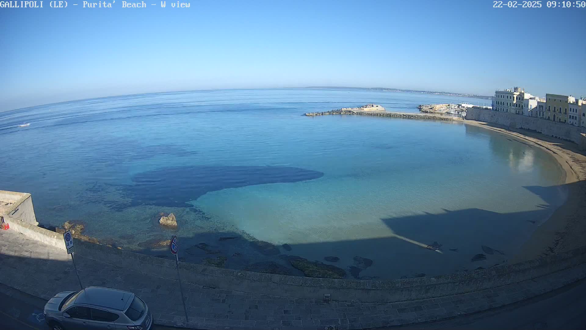 A clear, sunny day reveals a calm, turquoise bay curving along a sandy beach and stone buildings, with a car parked on a nearby road.