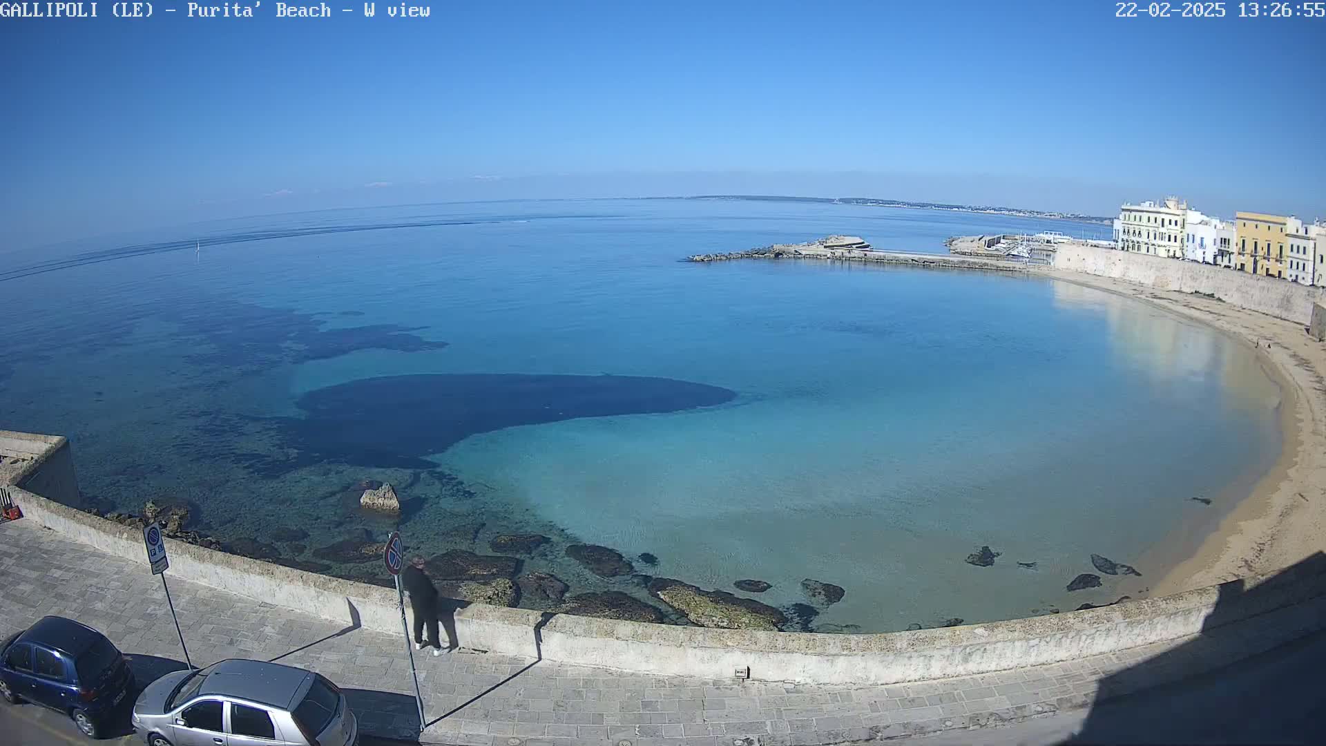 A clear, sunny day overlooks a calm, turquoise bay with a sandy beach and buildings along the shoreline.