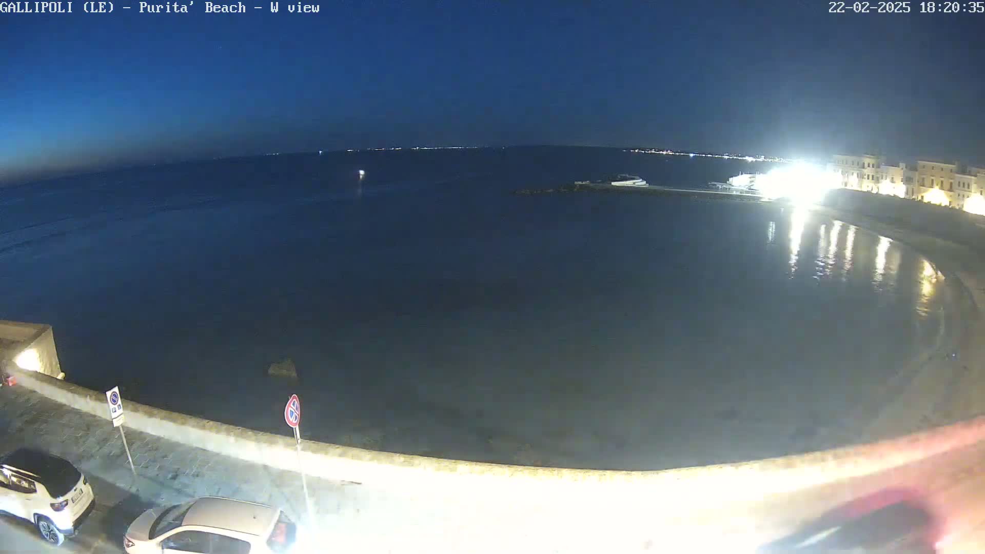 A nighttime view of a calm, dark-blue sea next to a low-lit, white stone waterfront with parked cars.