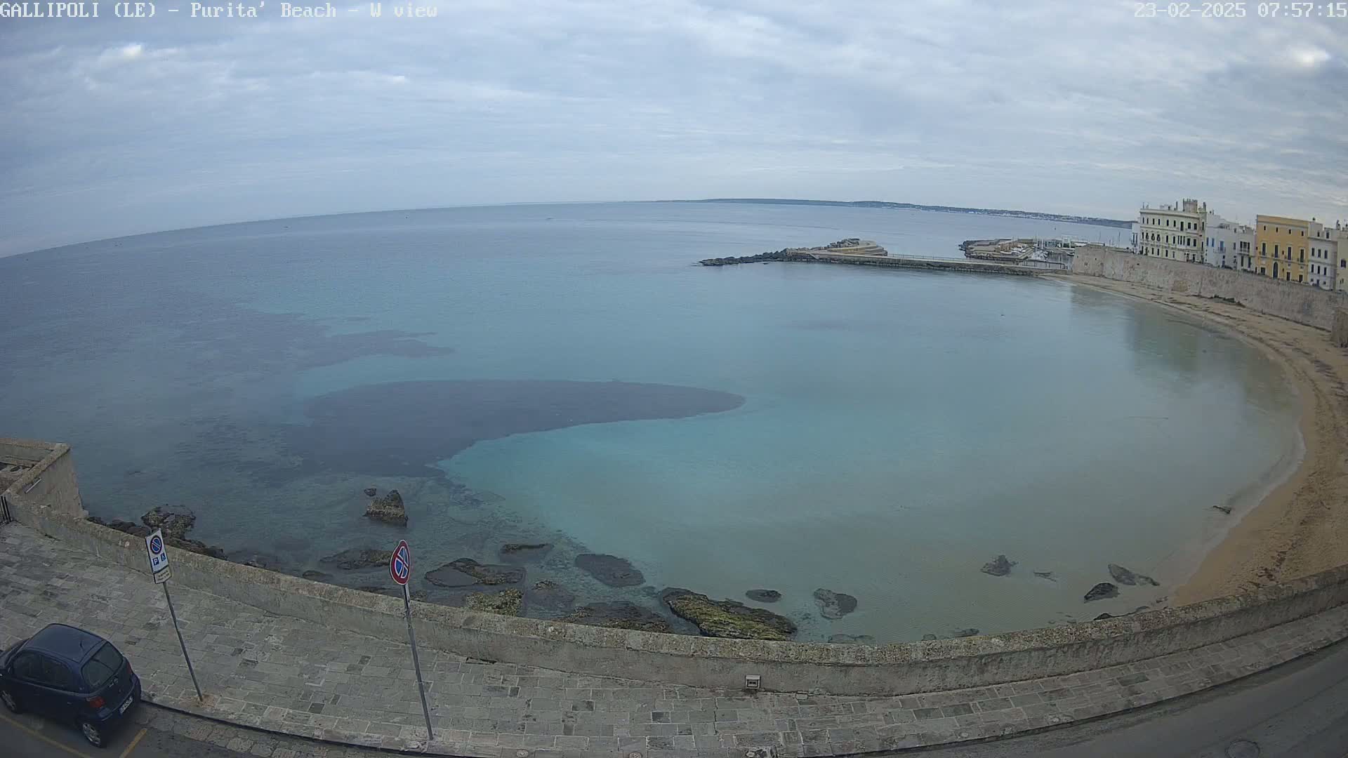 A beach with calm, light-blue water curves around a stone wall and buildings under an overcast sky.