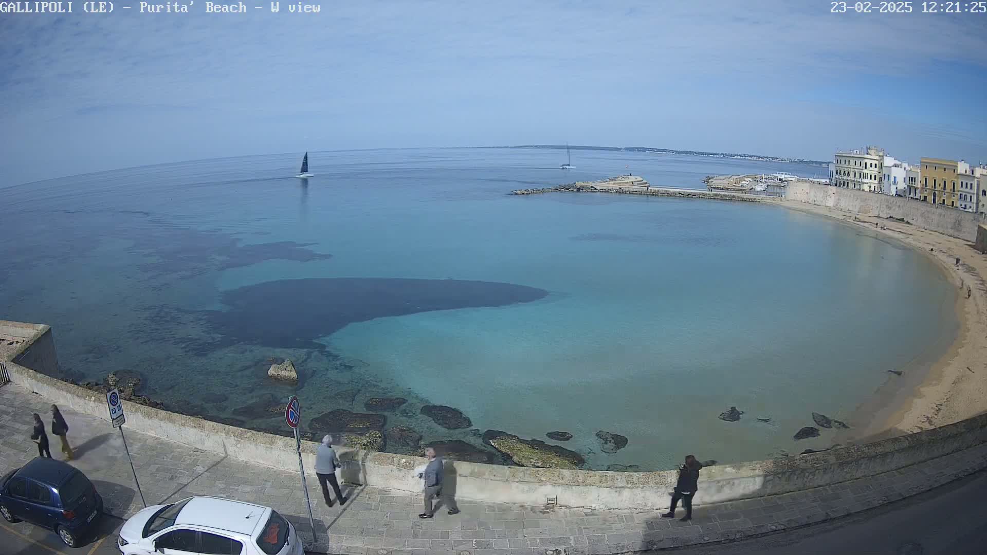 A clear, sunny day overlooks a calm, turquoise bay with a sandy beach, a low stone wall, several people walking, and a sailboat on the horizon.