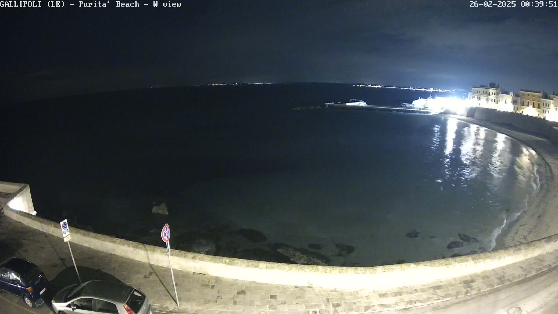 A nighttime view of a calm, dark bay curving along a stone wall with buildings and two cars parked on the street.