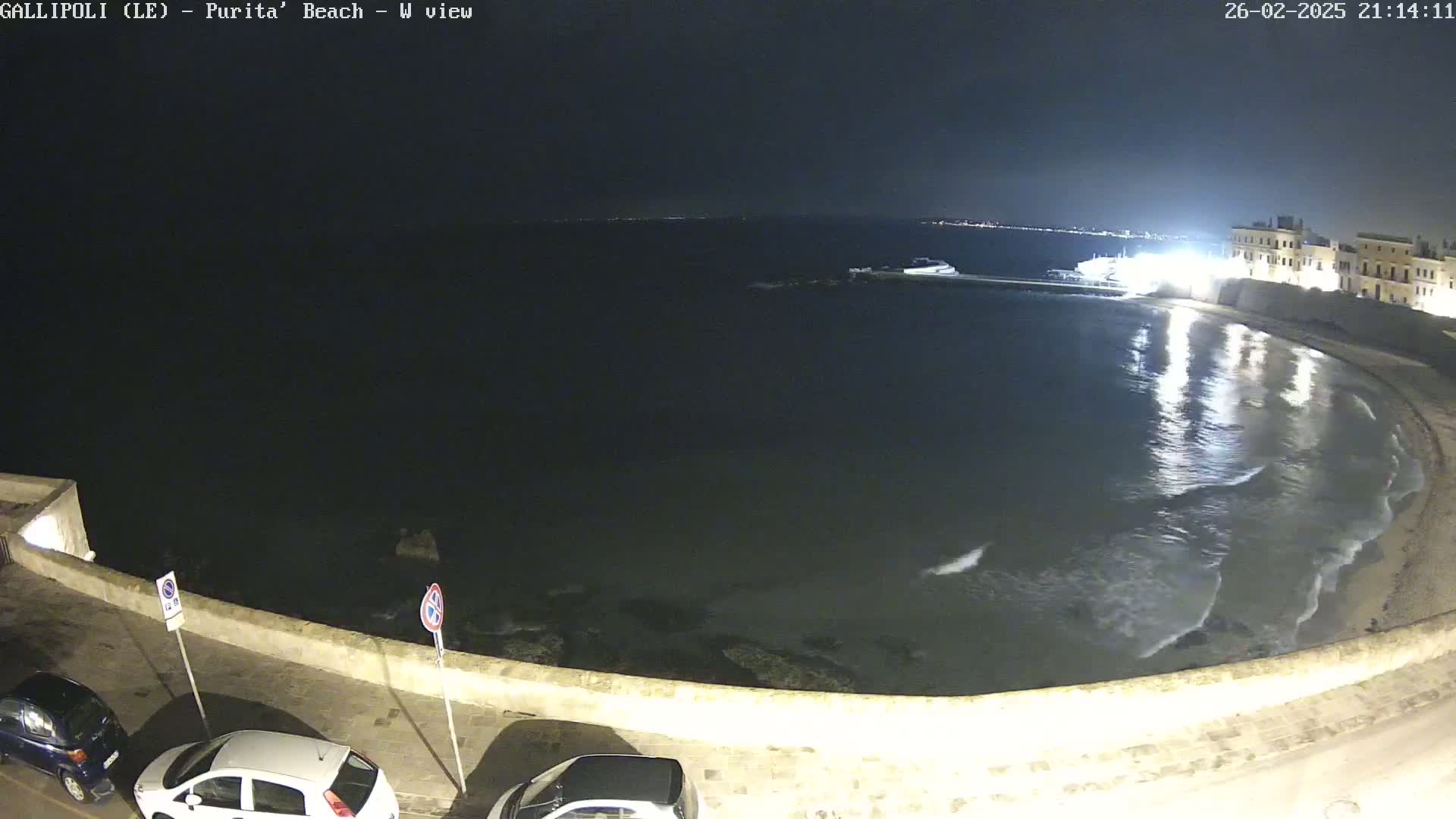A nighttime view of a calm bay with gentle waves lapping a crescent-shaped beach, bordered by a low stone wall and a few parked cars.