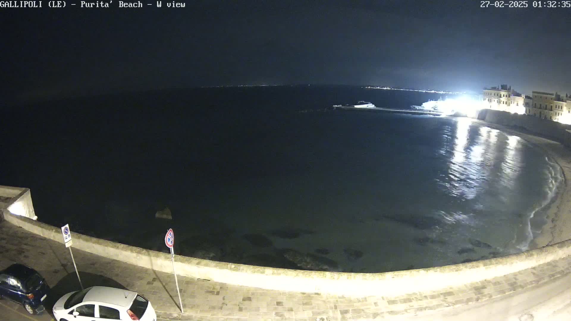 A nighttime, seaside view shows a calm, dark sea lapping a gently curving shore next to a paved walkway with parked cars.