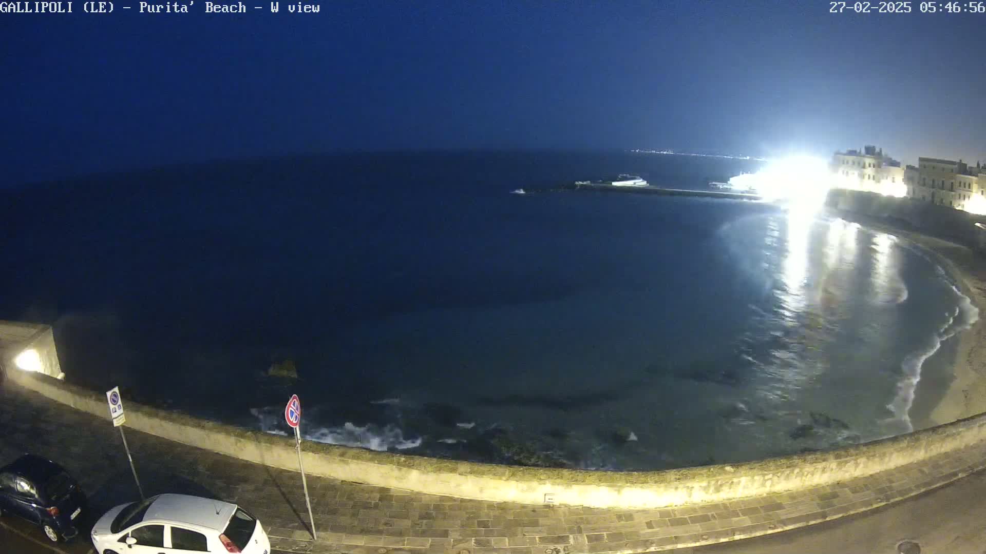 A nighttime view of a calm, dark-blue sea lapping gently against a stone wall, with a few cars parked nearby and buildings illuminated in the background.