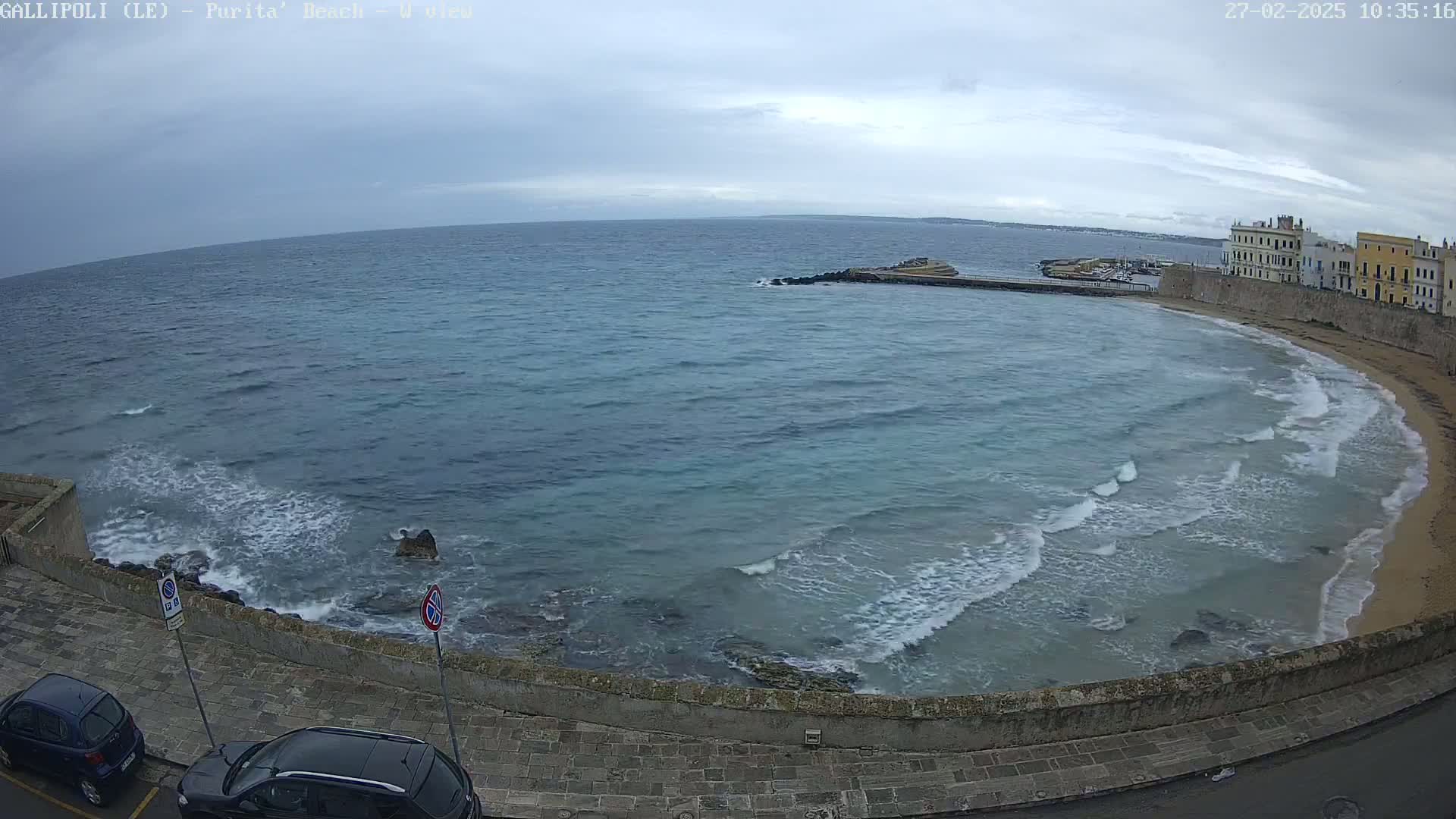 A partly cloudy day shows a curving shoreline with a sandy beach and buildings, a calm sea with small waves lapping against the shore, and two cars parked along a stone wall.