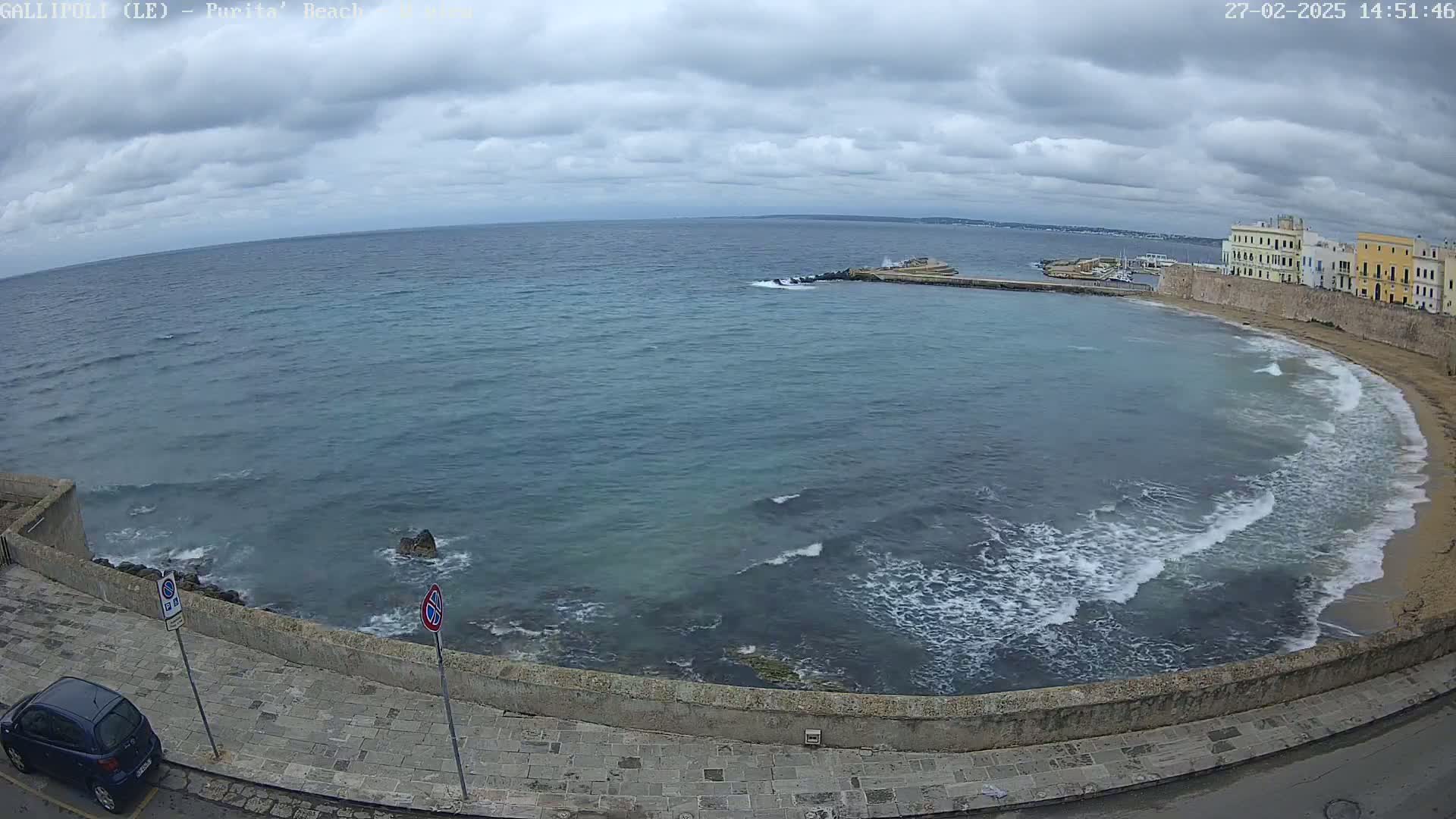 A coastal scene shows a partly cloudy sky over a calm sea lapping a sandy beach next to a stone wall and buildings.
