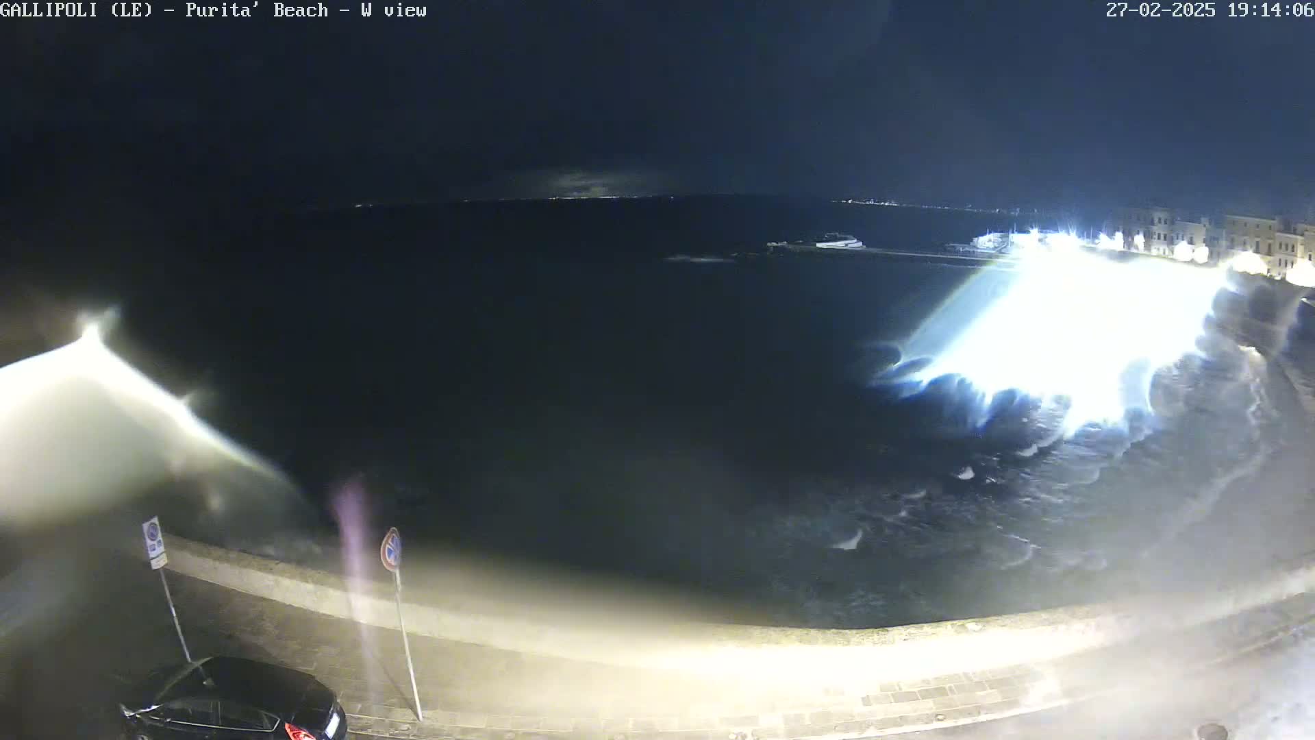 At night, large waves crash against a seawall next to a parked car under stormy conditions.