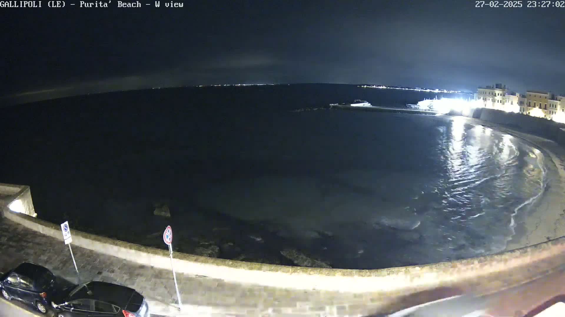 A nighttime view of a calm, dark ocean bay next to a stone wall and a street with parked cars, under a clear sky.
