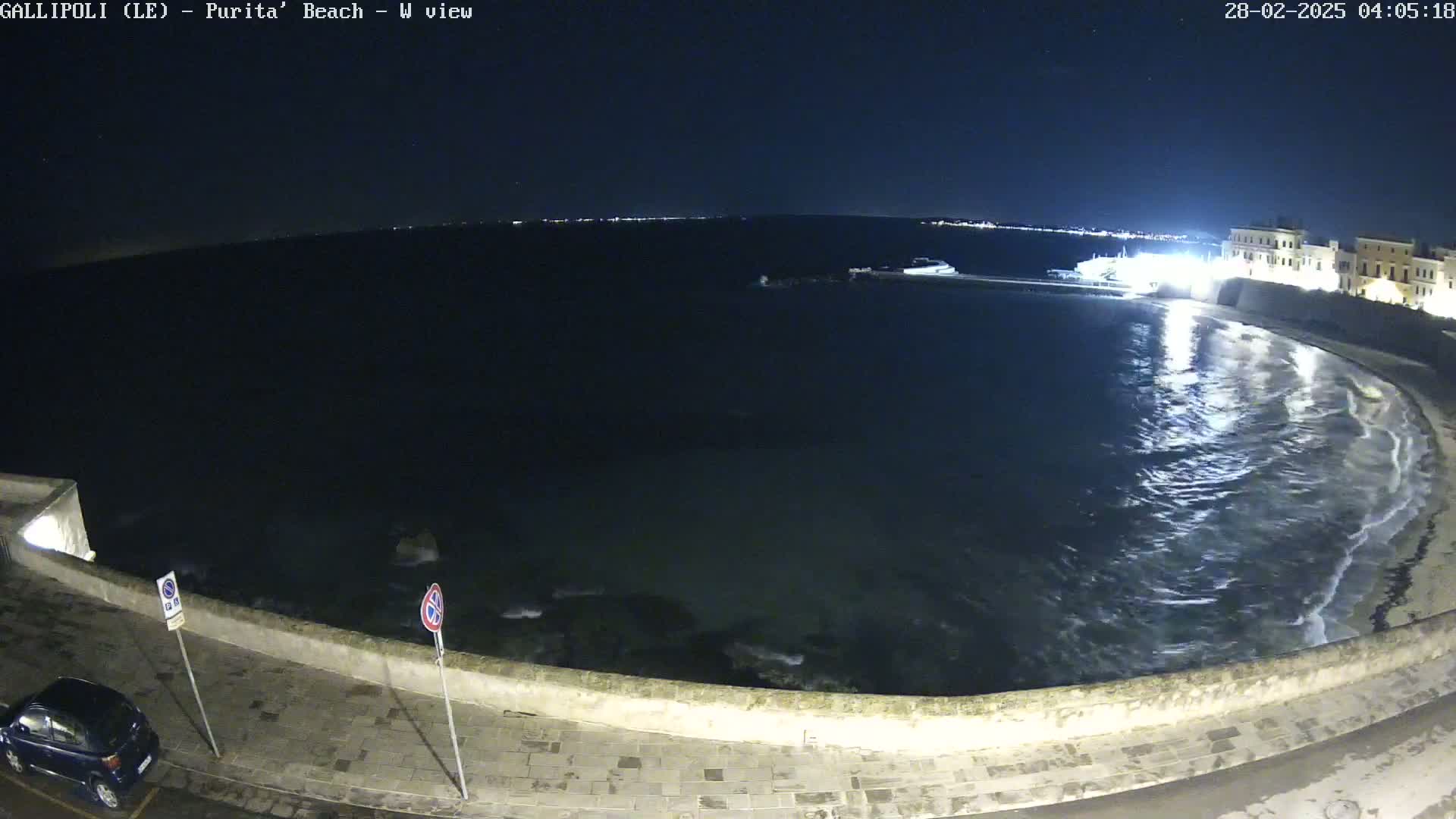 A nighttime, seaside view shows gently lapping waves on a dark, calm sea next to a low stone wall, a paved road, and a parked car.