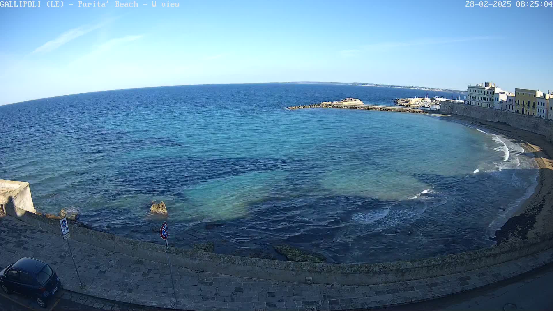 A clear, sunny day shows a calm bay with light waves lapping a sandy shore next to a stone walkway and buildings.