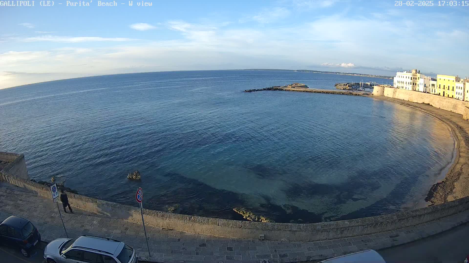 A partly sunny day overlooks a calm, clear bay curving around a stone wall and buildings, with a few people and parked cars visible along the wall.