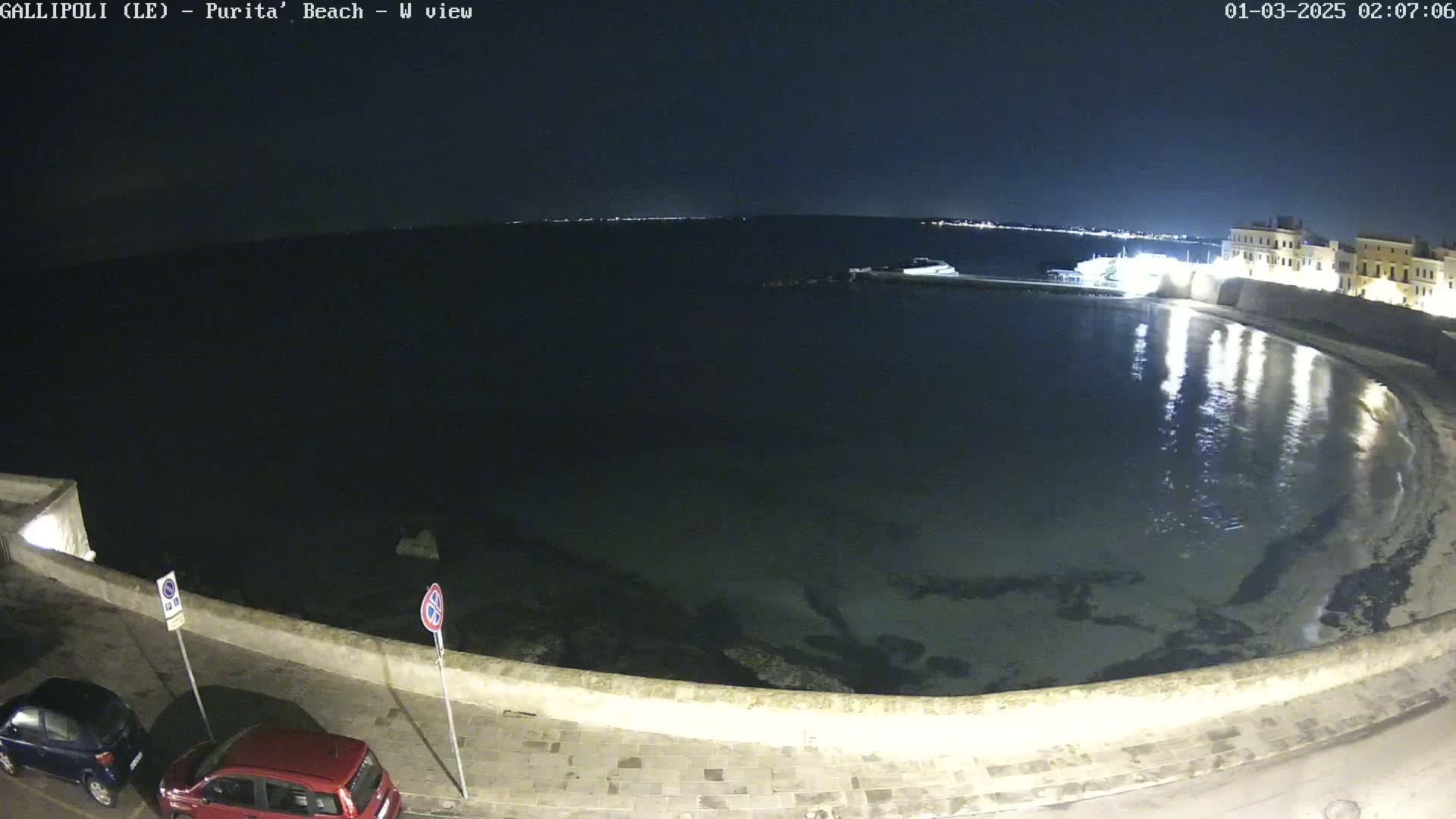 A nighttime view of a calm, dark body of water curving along a stone wall, with a few cars parked along the street next to it and distant lights visible across the water.