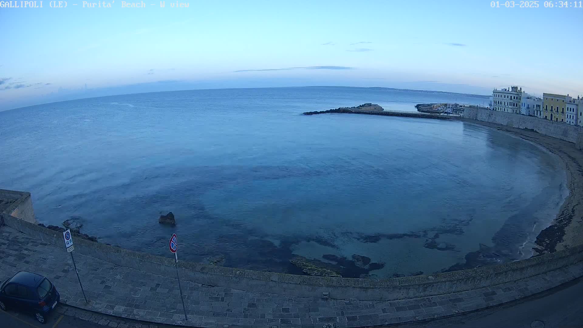 A calm, blue-toned sea laps against a stone embankment near buildings under a clear, pre-dawn sky.