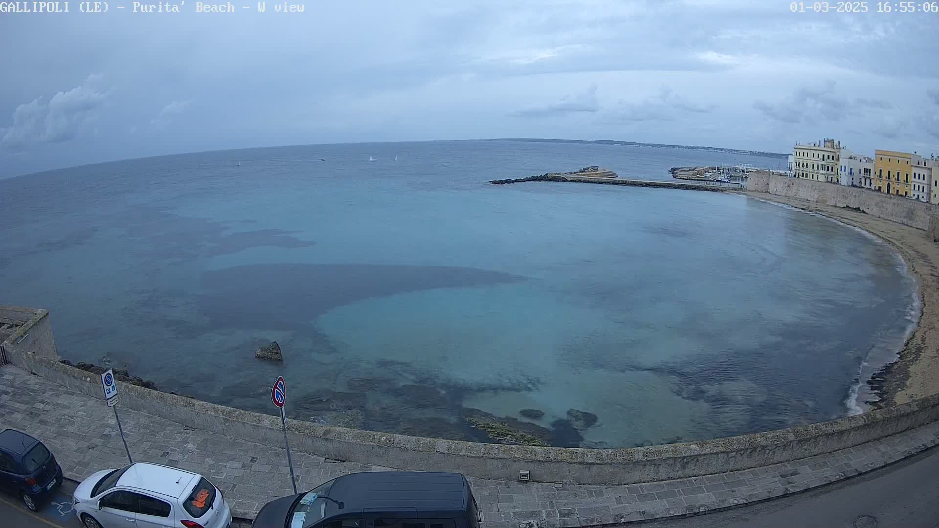 A partly cloudy day overlooks a calm, teal bay curving along a stone wall next to a coastal town with parked cars.