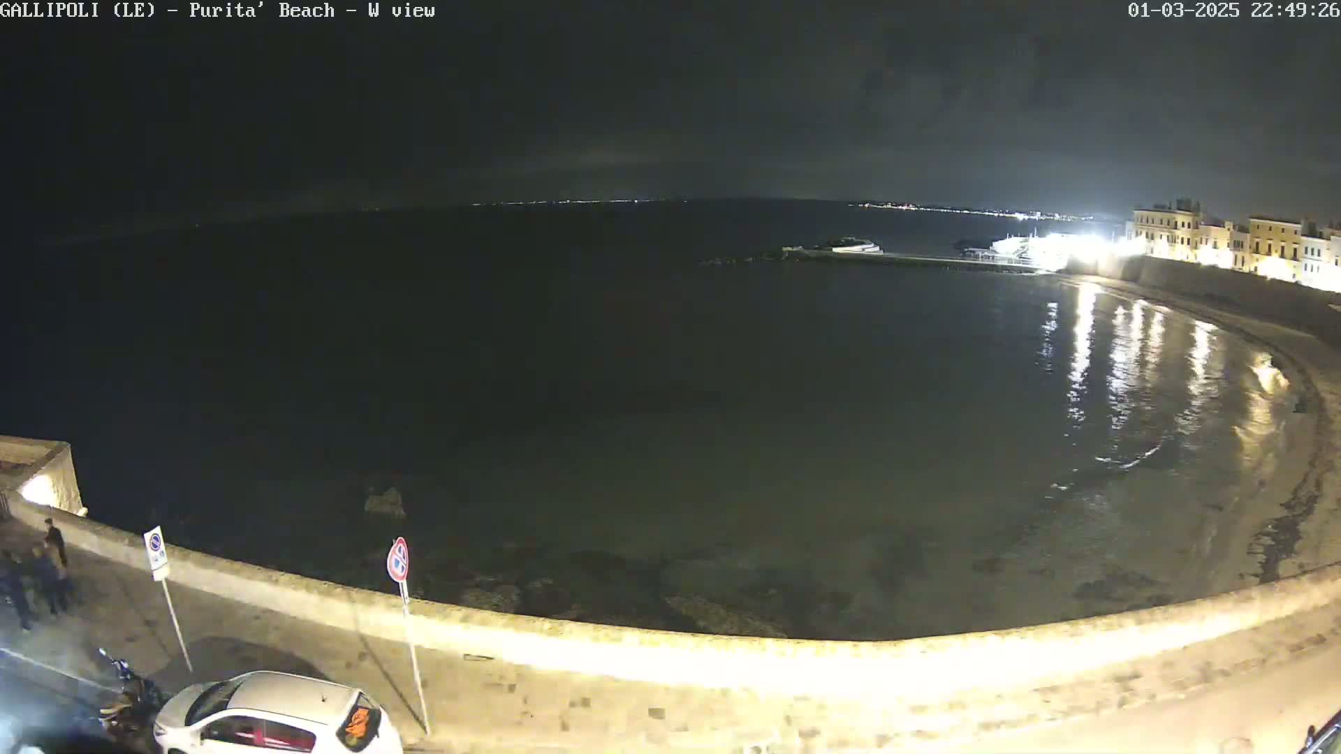 A nighttime view of a calm bay, partially enclosed by a stone wall, with a few cars parked along the wall.