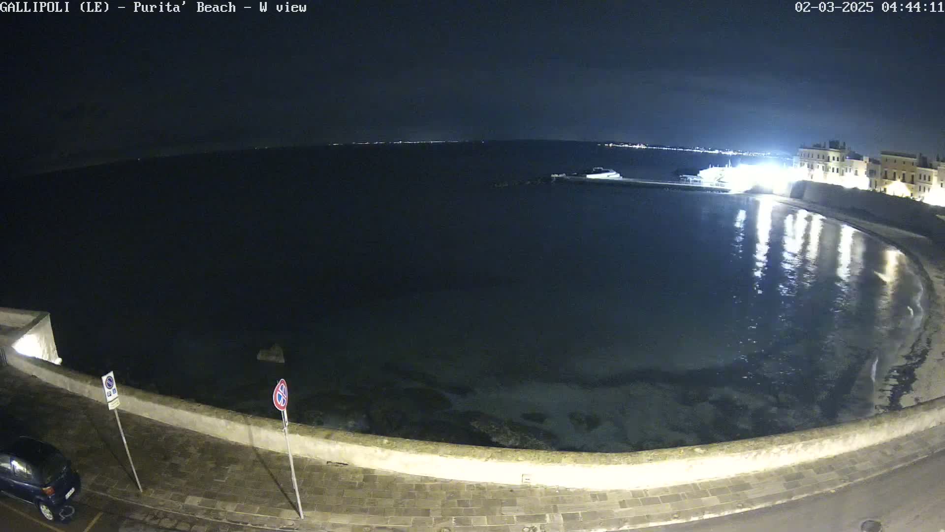 A nighttime view of a calm, dark body of water curving along a lit stone wall with a car parked nearby under a clear, dark sky.