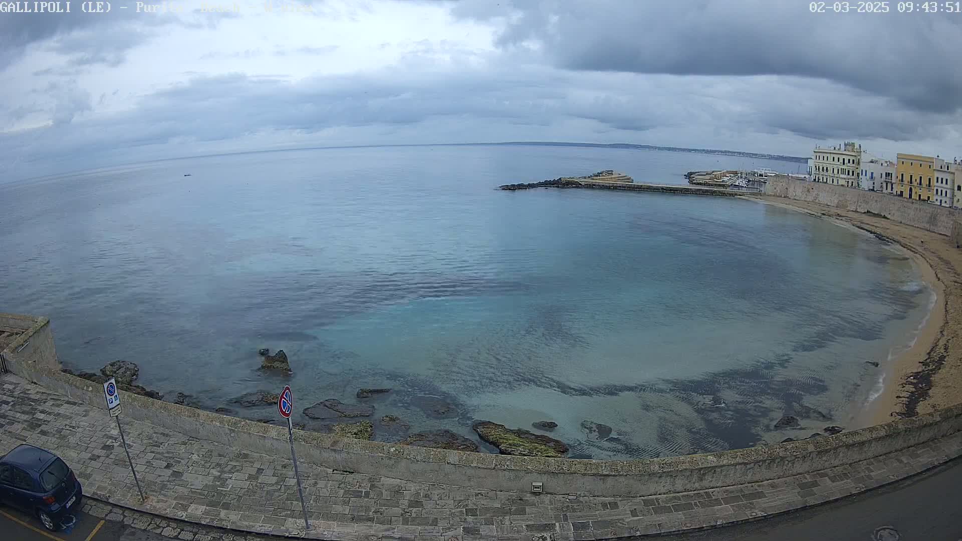A coastal scene under a cloudy sky shows a calm, shallow bay with a sandy beach, stone walls, and buildings along the shore.