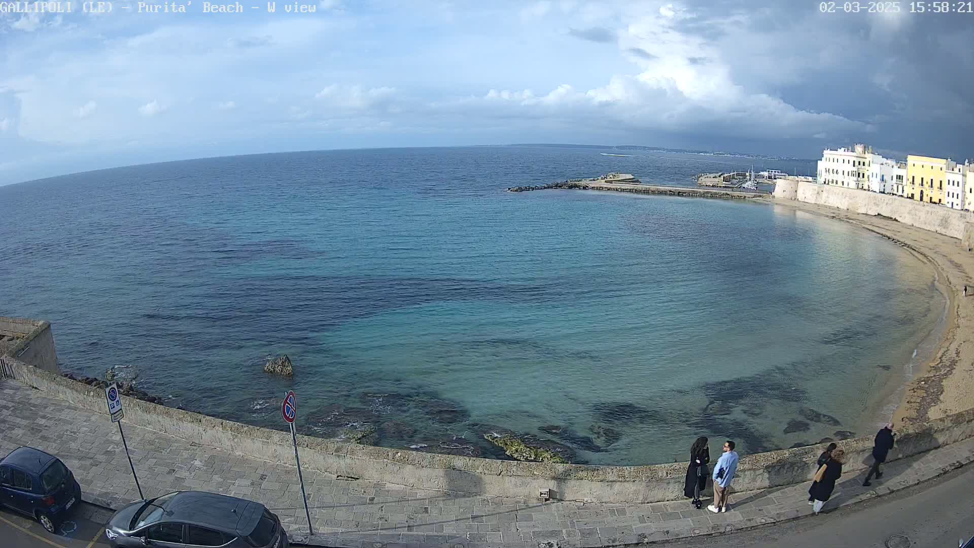 A partly cloudy day reveals a curved beach with turquoise water, a stone wall, and several people strolling along a paved walkway.