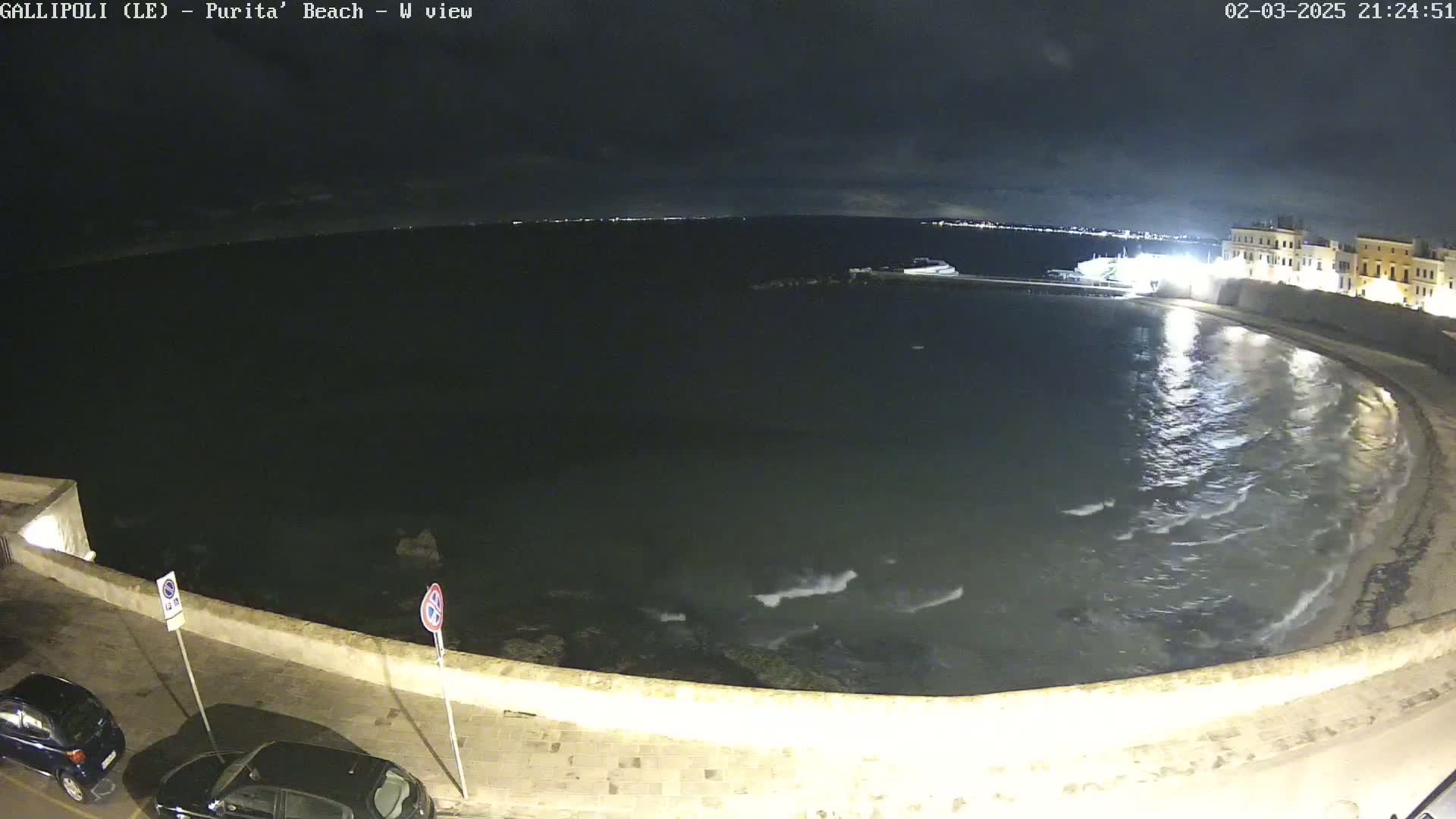 A nighttime view of a calm, dark-colored sea lapping gently against a curved shoreline, with a town's lights visible in the distance under a cloudy sky.