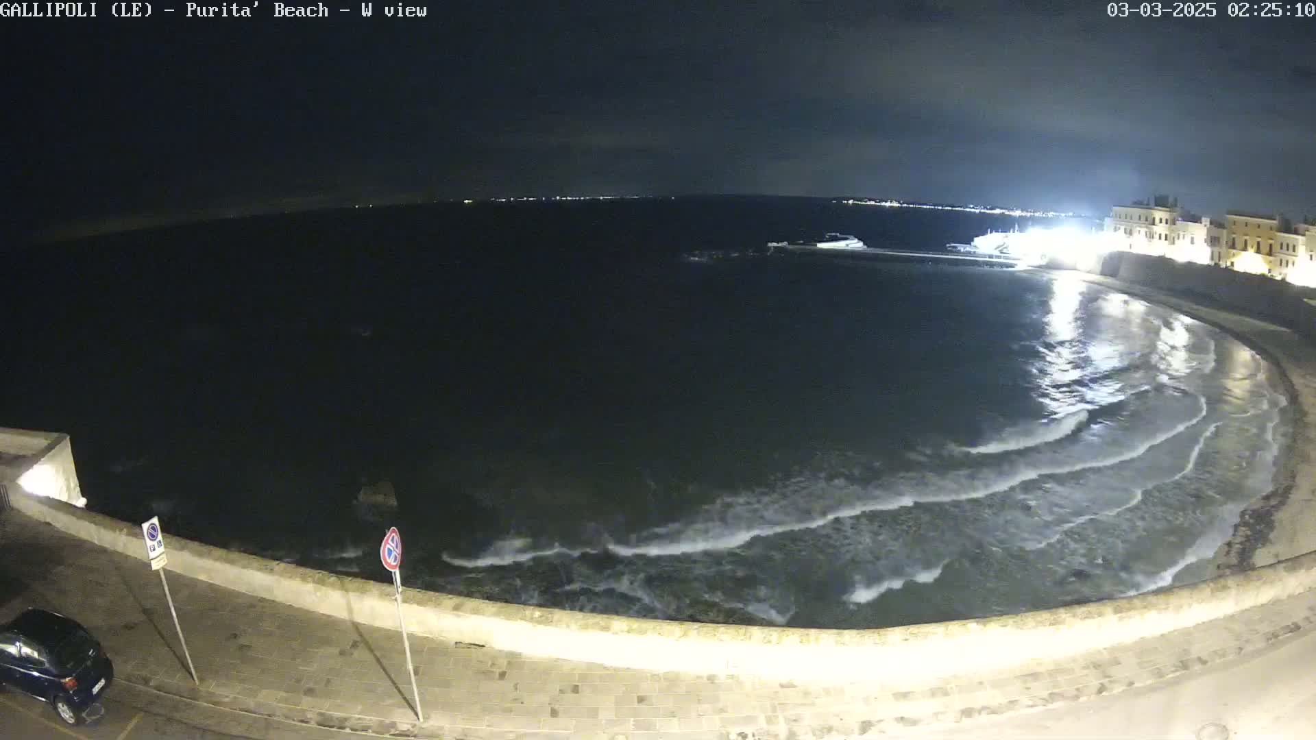 A nighttime view of a calm sea lapping gently at a stone wall next to a paved area, with a building and lights visible in the distance.