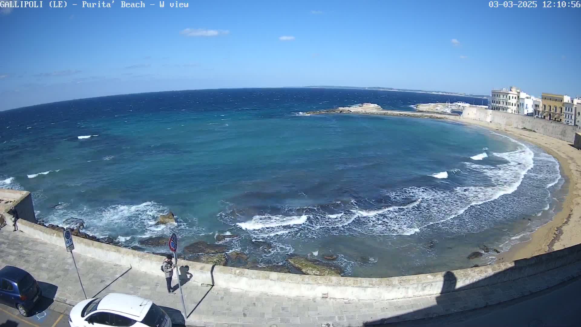 A partly sunny day shows a curved bay with turquoise water lapping a sandy beach, bordered by a low stone wall and buildings, with small waves breaking near the shore.