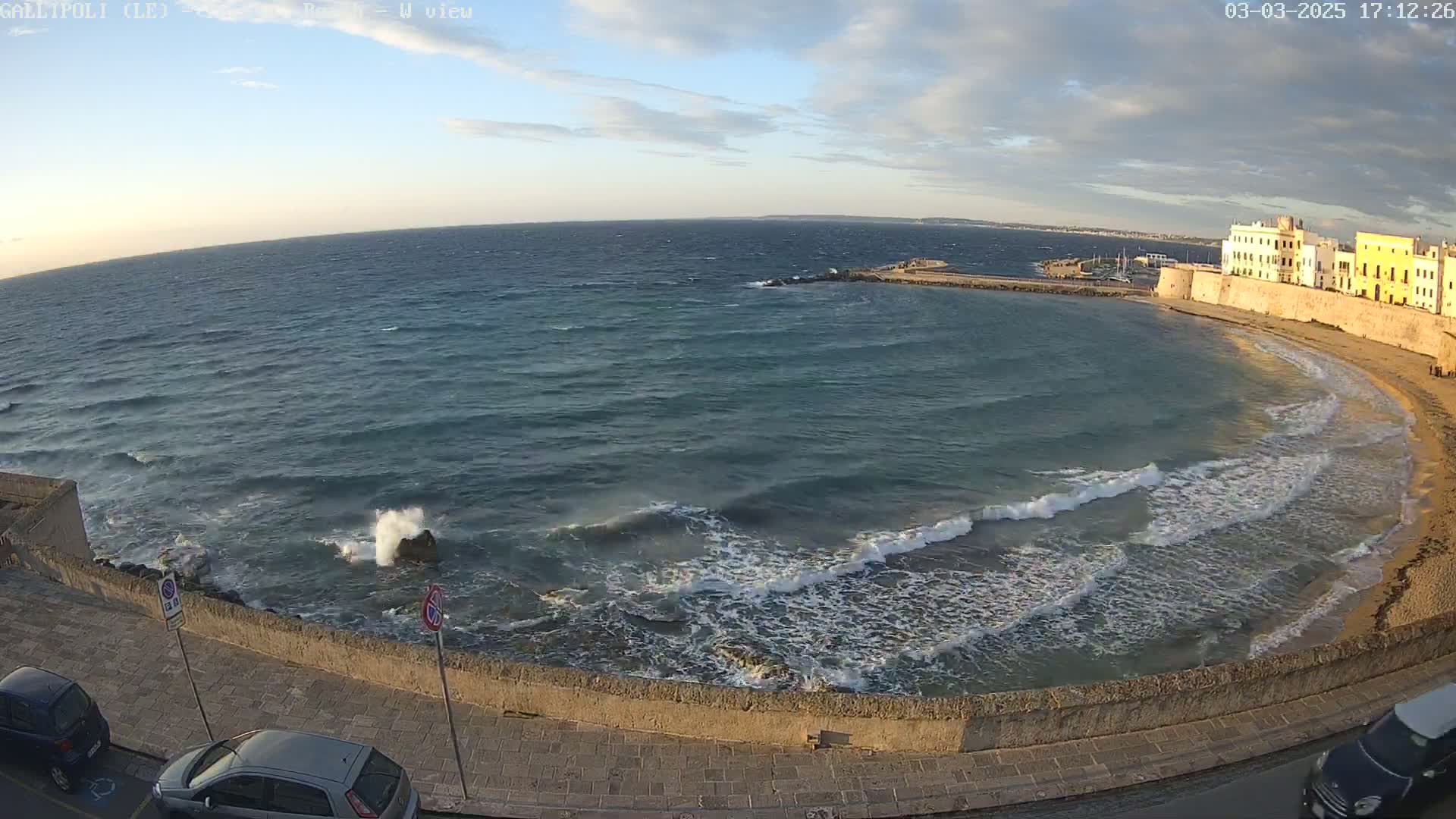 A partly cloudy sunset over a wavy sea laps against a sandy beach and stone wall near a town.