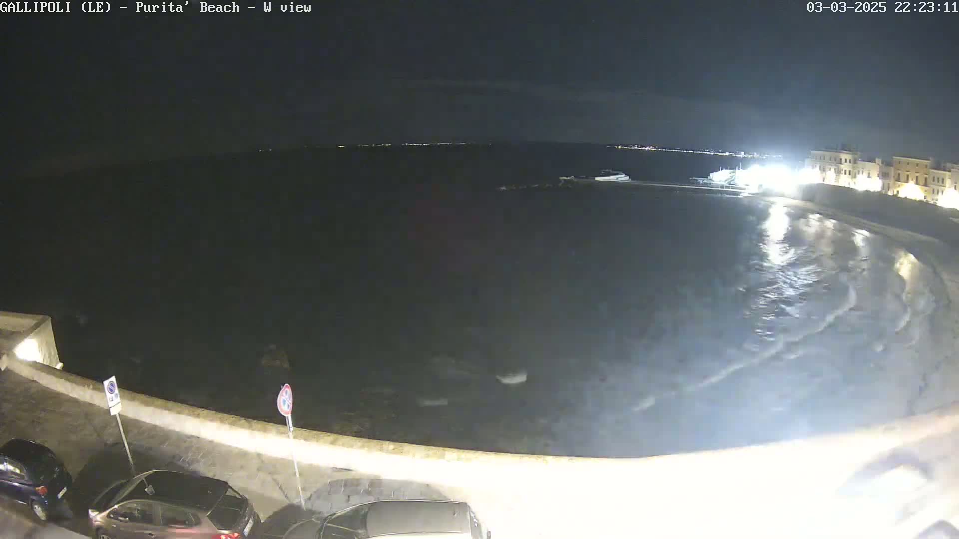 A nighttime view of a dark, wavy ocean lapping against a stone wall, with a few parked cars visible along the wall.