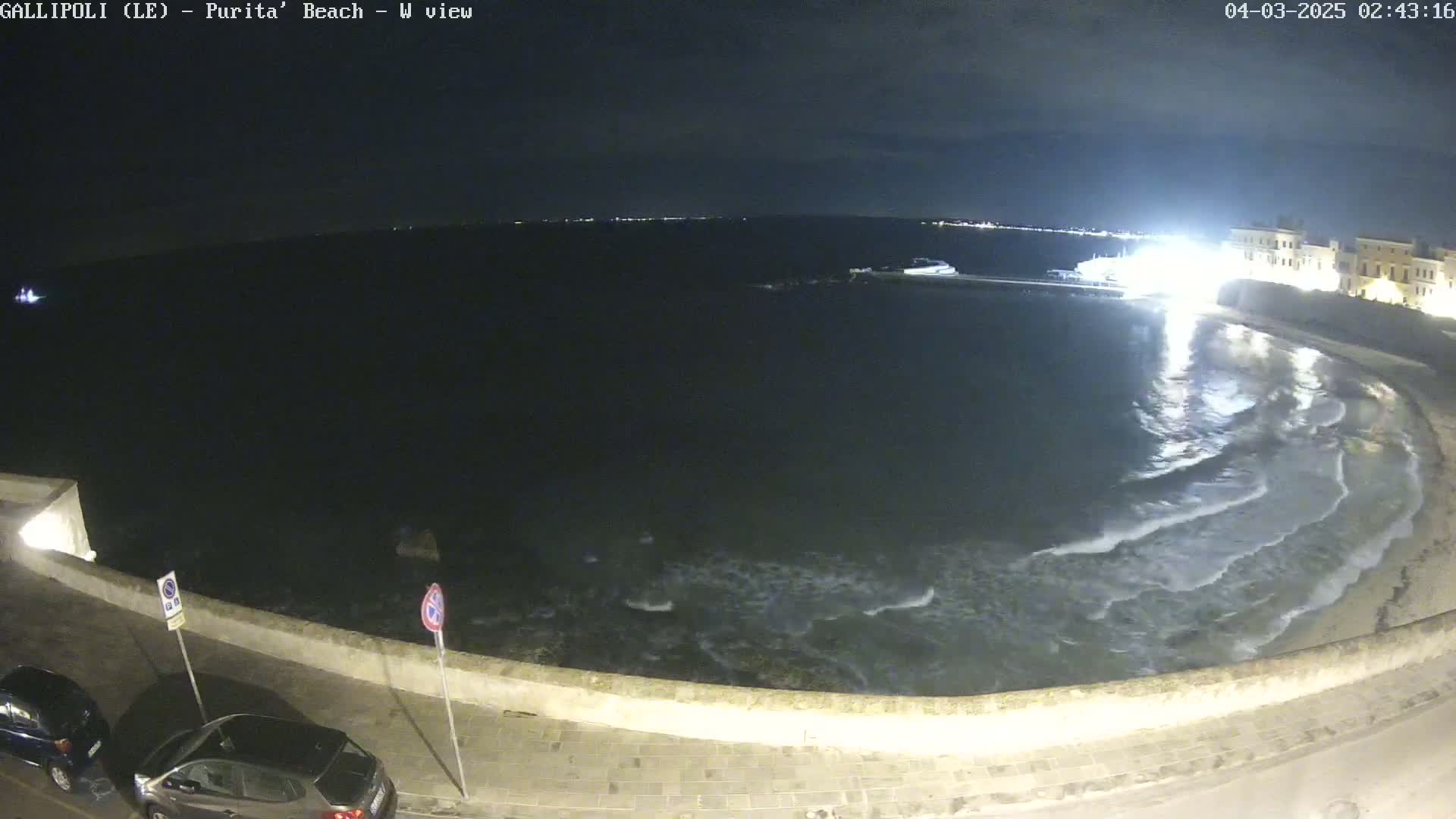 A nighttime view of a calm sea lapping gently against a stone wall bordering a paved area with parked cars.
