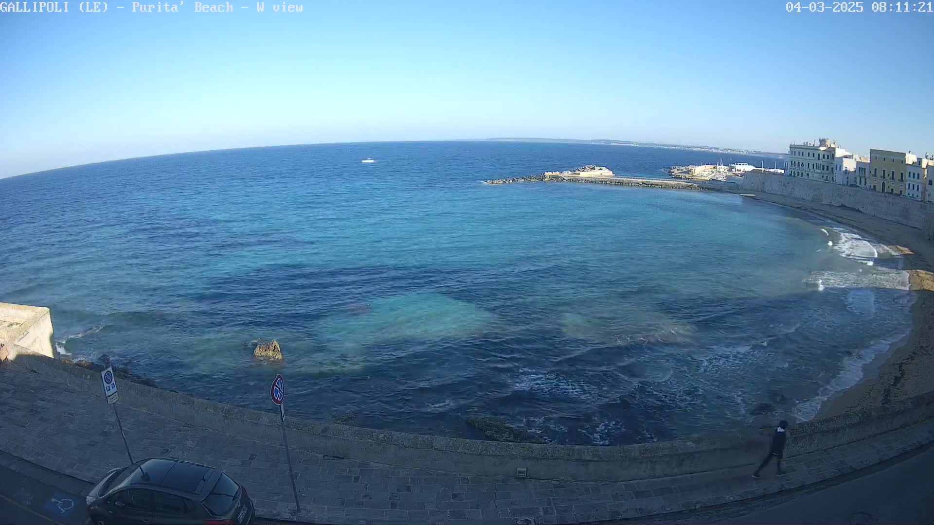 A clear, sunny day shows a turquoise ocean lapping against a sandy beach next to a stone wall and buildings, with a person walking along the wall.