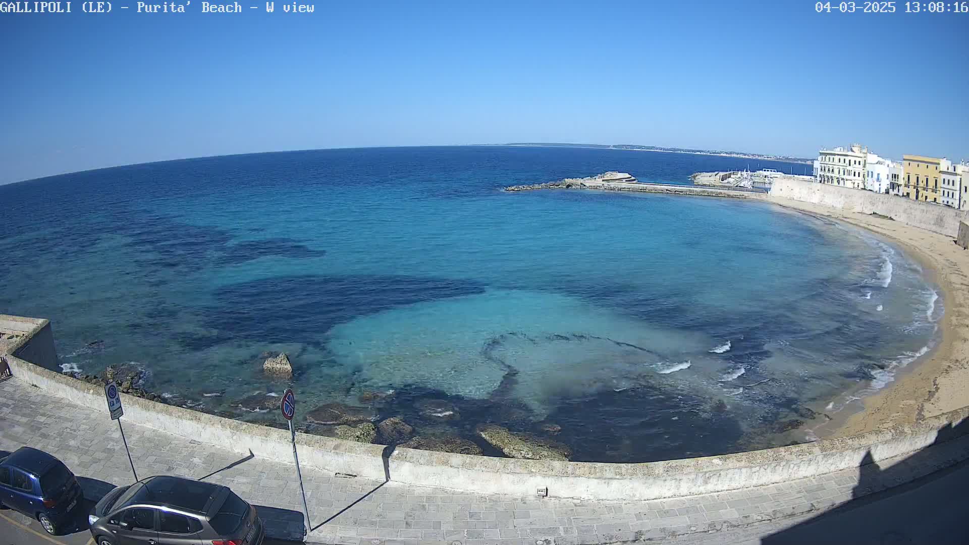 A clear, sunny day reveals a picturesque coastal scene with turquoise water lapping a sandy beach, bordered by a stone wall and buildings.